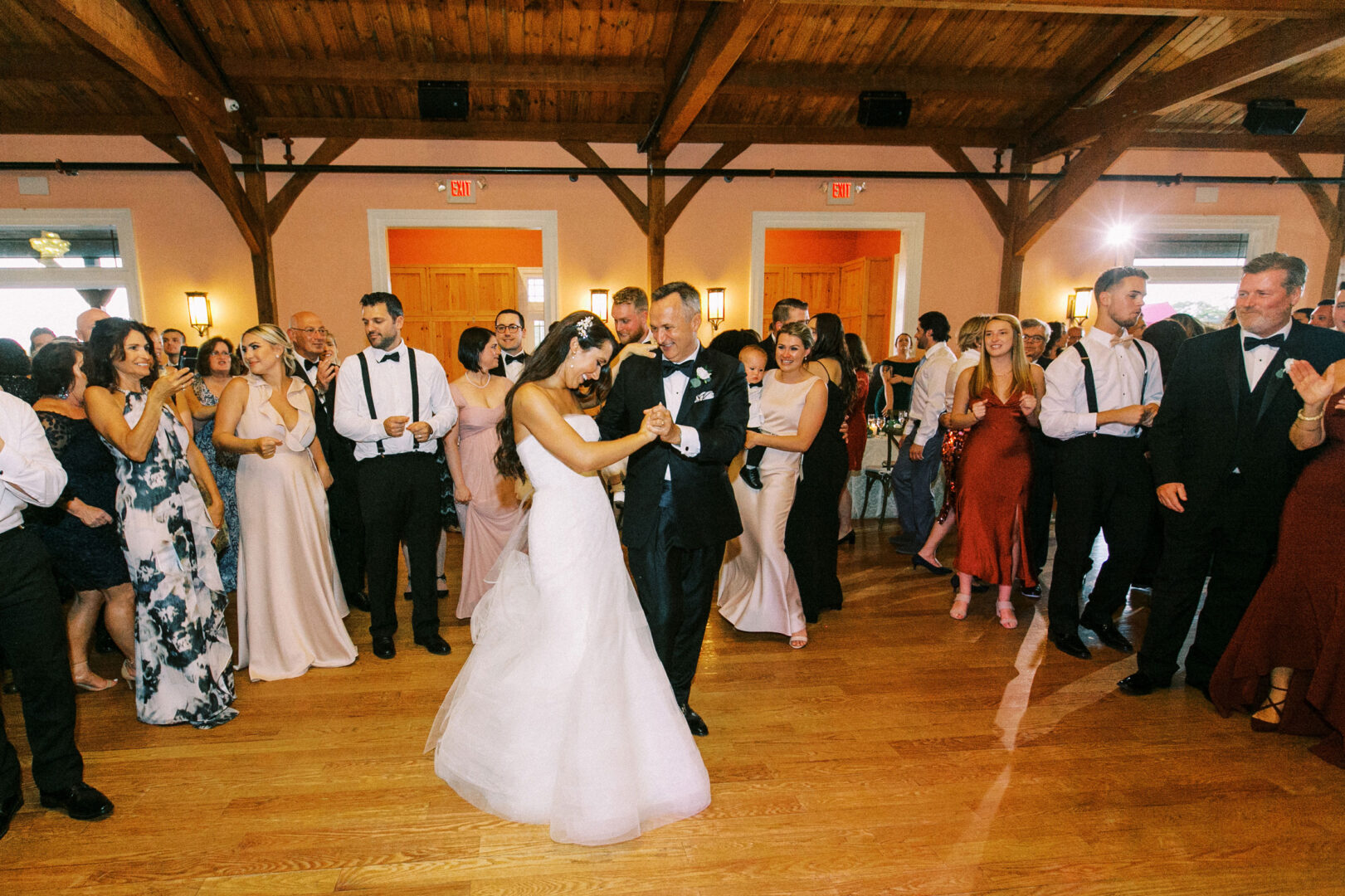 At a Willow Creek Winery wedding, a bride and groom dance surrounded by guests on a wooden dance floor in a warmly lit reception hall.