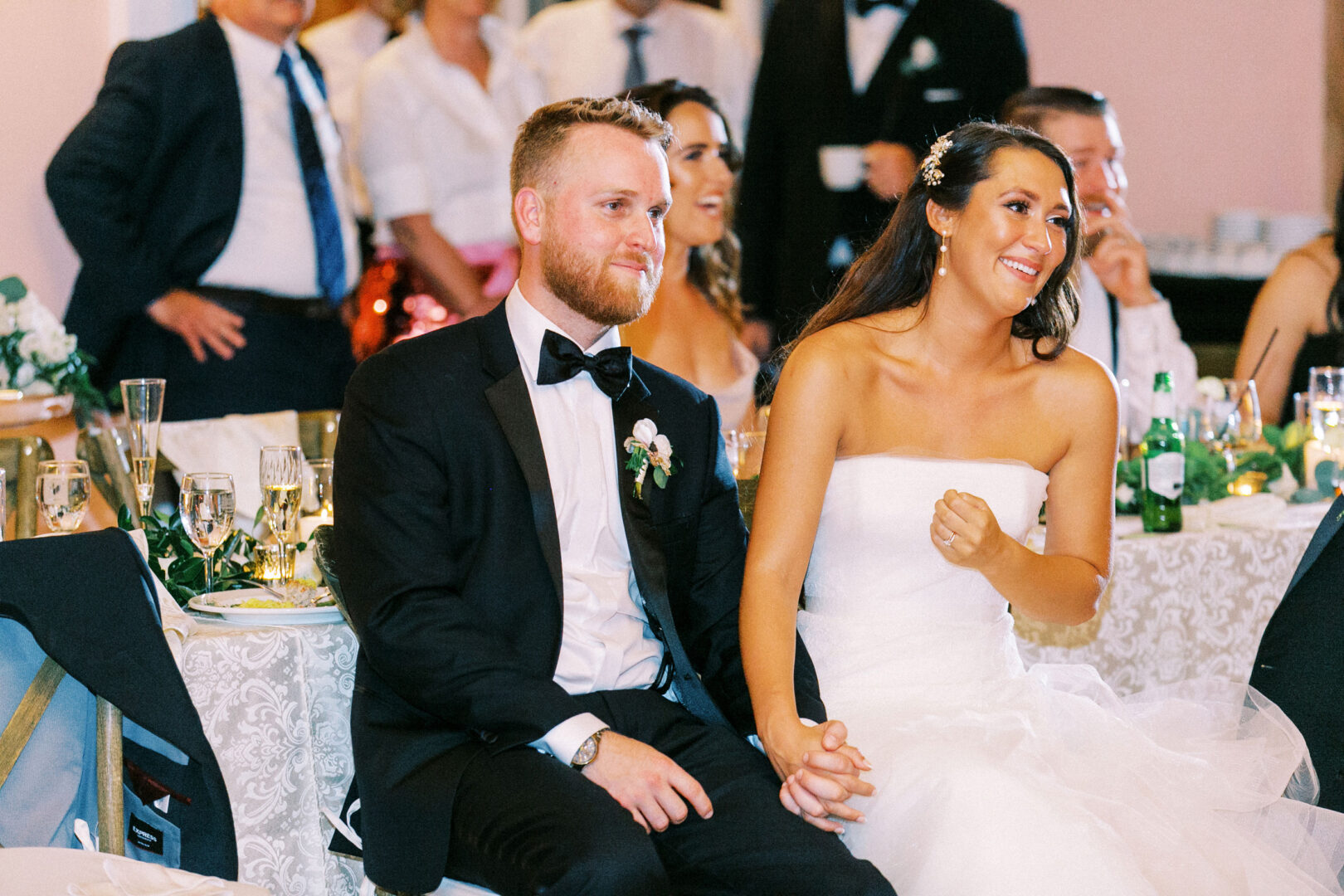 At the Willow Creek Winery Wedding, a bride and groom sit at a table, smiling and holding hands, surrounded by guests at a joyful reception.