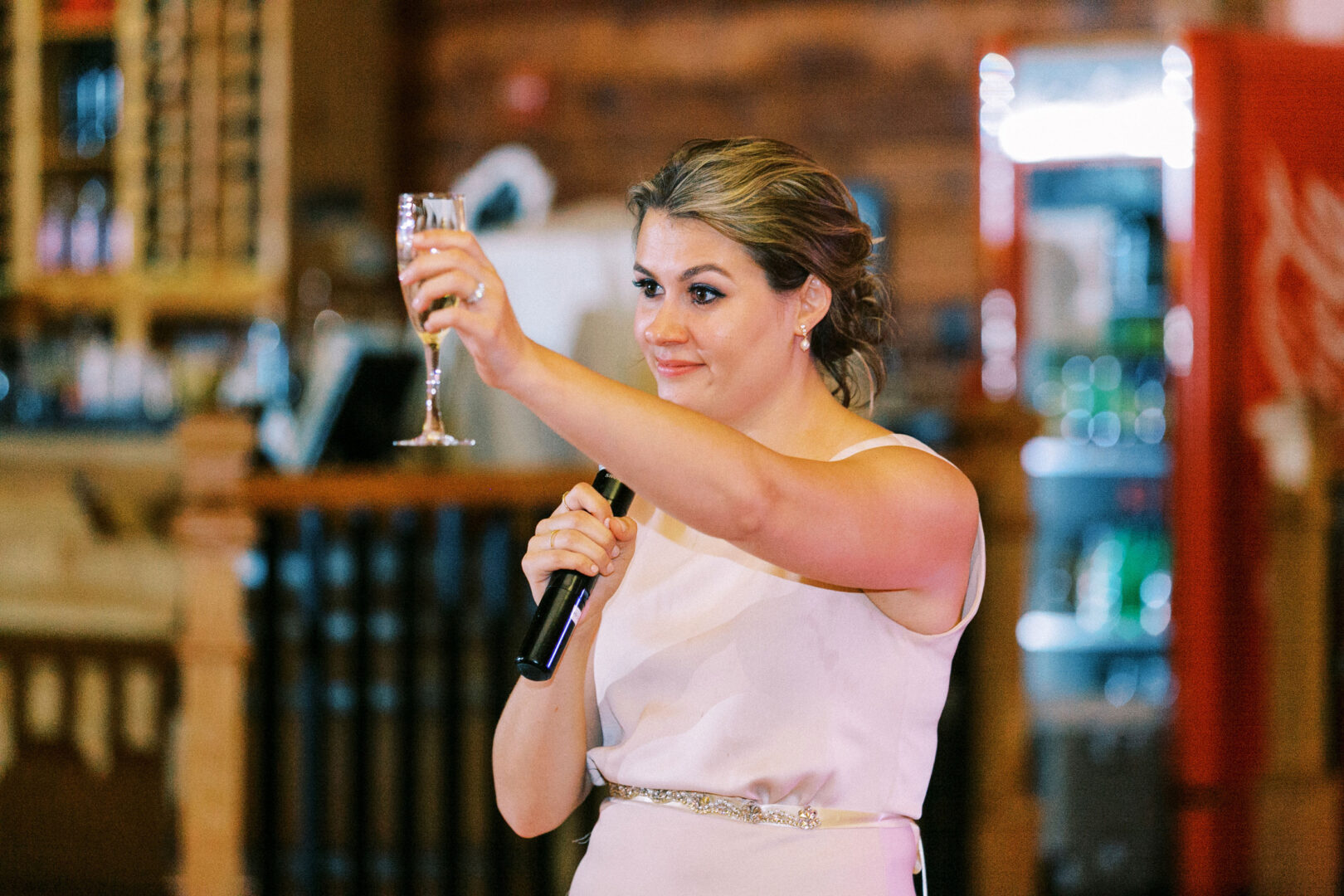 A woman in a white dress holds a microphone and raises a glass in a toast, capturing the joyous spirit of a Willow Creek Winery wedding inside the warmly lit room.