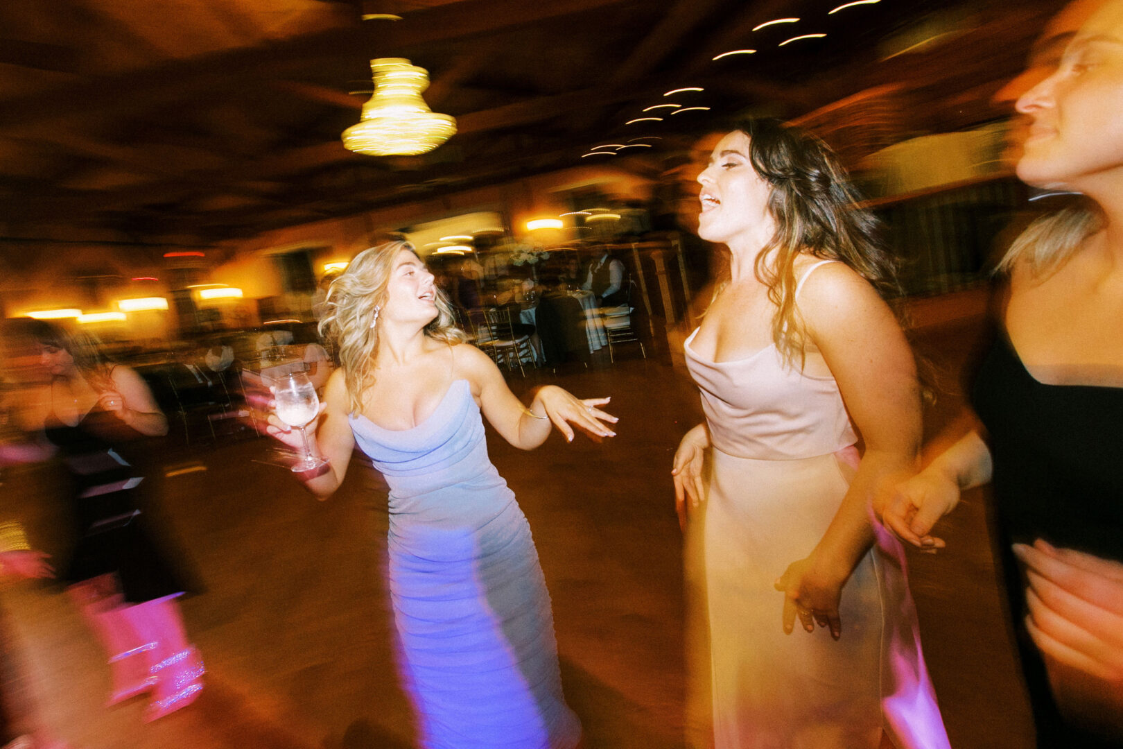 At a Willow Creek Winery wedding, three women in formal dresses dance energetically in the warmly lit venue. One holds a drink, while the others gesture with their hands against a backdrop of blurred lights and mingling guests.