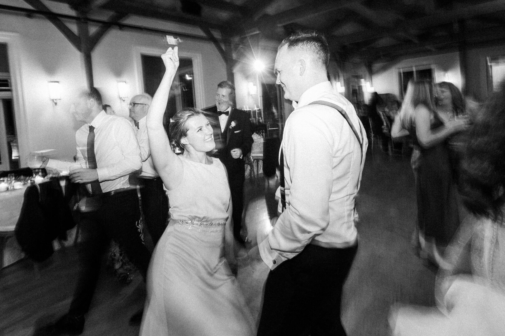 A black and white photo captures a lively dance floor at a Willow Creek Winery wedding. A woman in a dress enthusiastically dances with a man in a shirt and suspenders, as other guests join the celebration in the background.