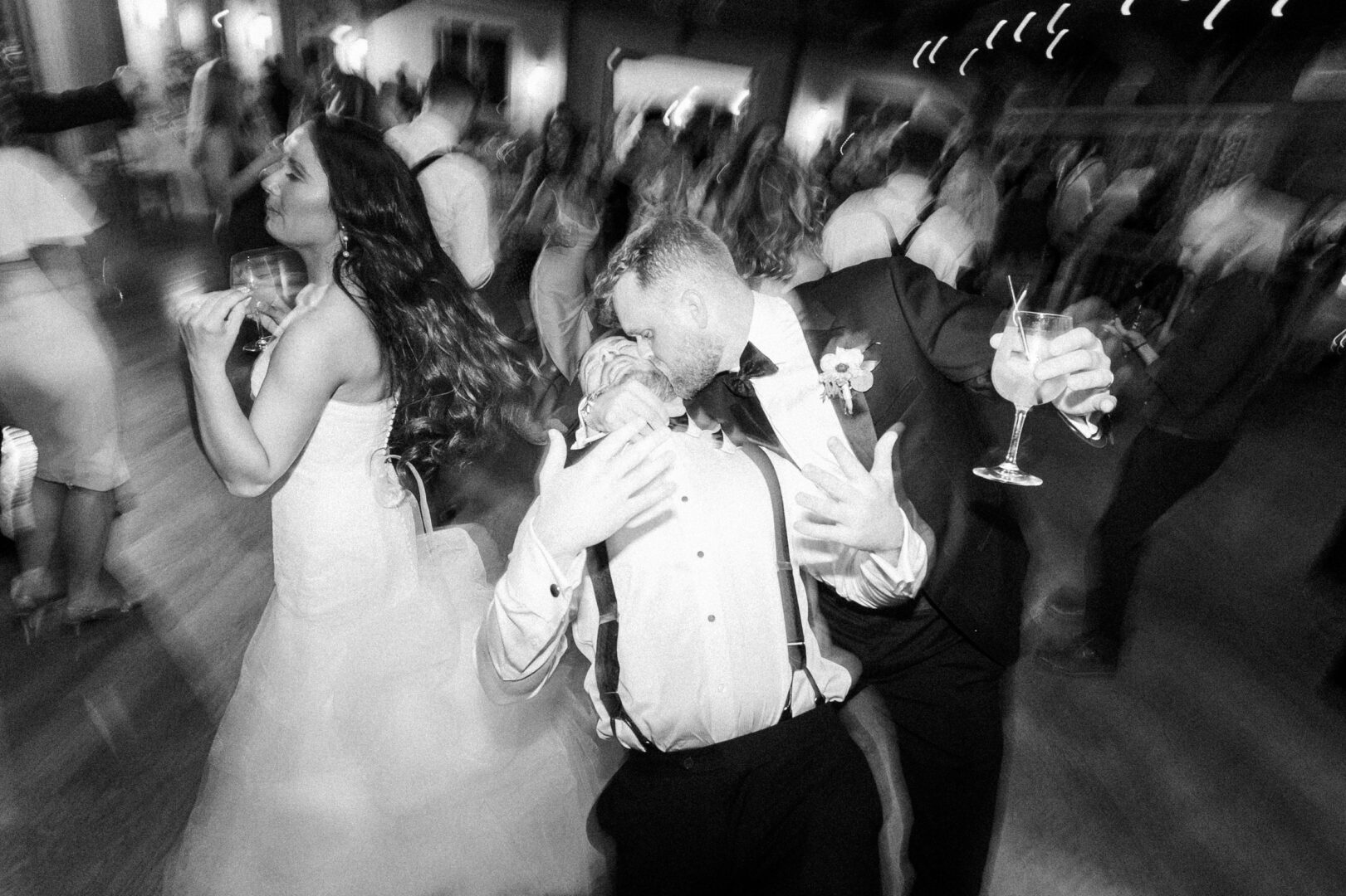 People celebrating at a lively Willow Creek Winery wedding reception; a man in a bow tie holding a drink, and a woman in a wedding dress smiling and dancing. The image is blurred, conveying motion.