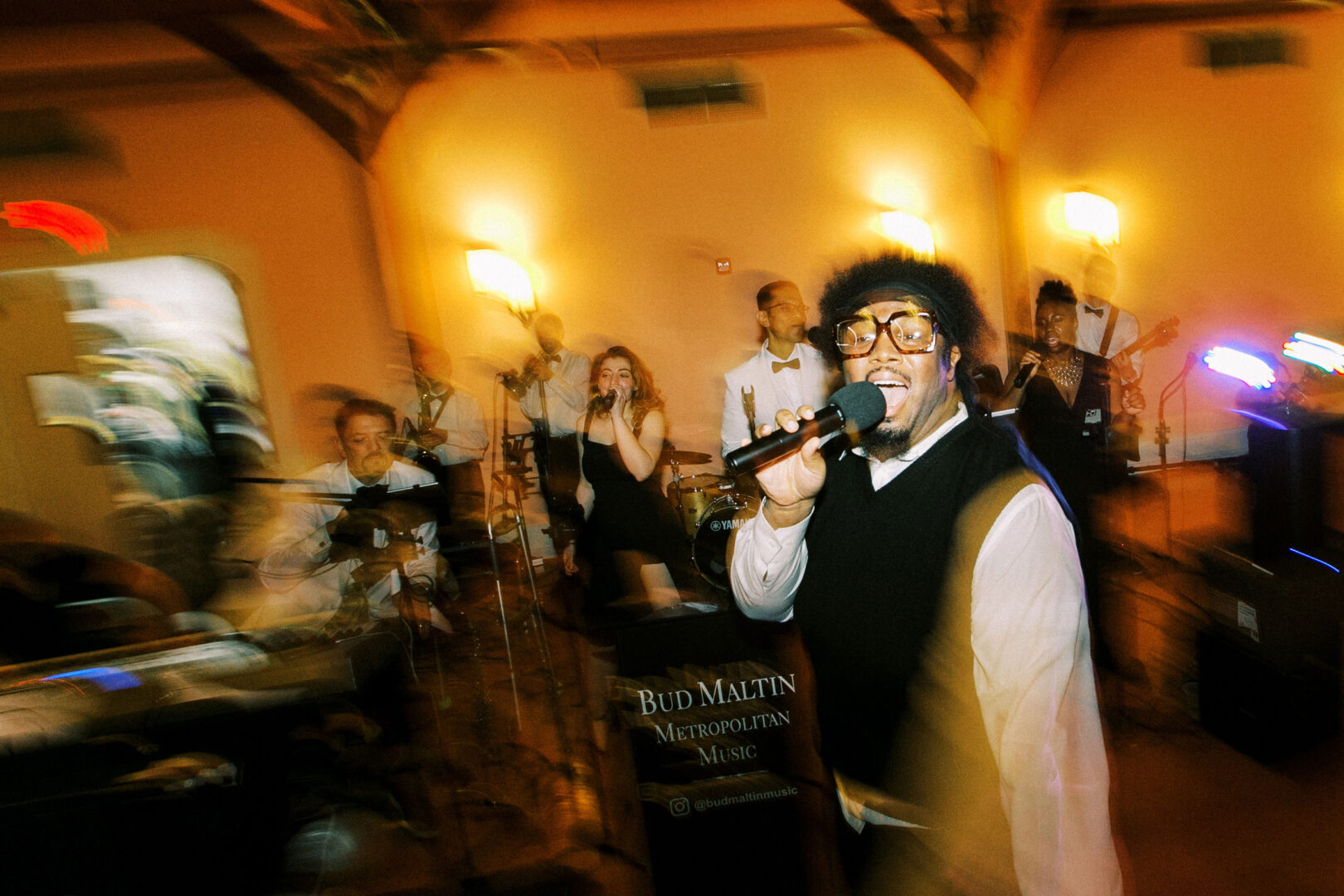 Amid the warmly lit room of the Willow Creek Winery Wedding, a band performs energetically on stage, with a singer in the foreground holding a microphone, surrounded by musicians playing instruments.