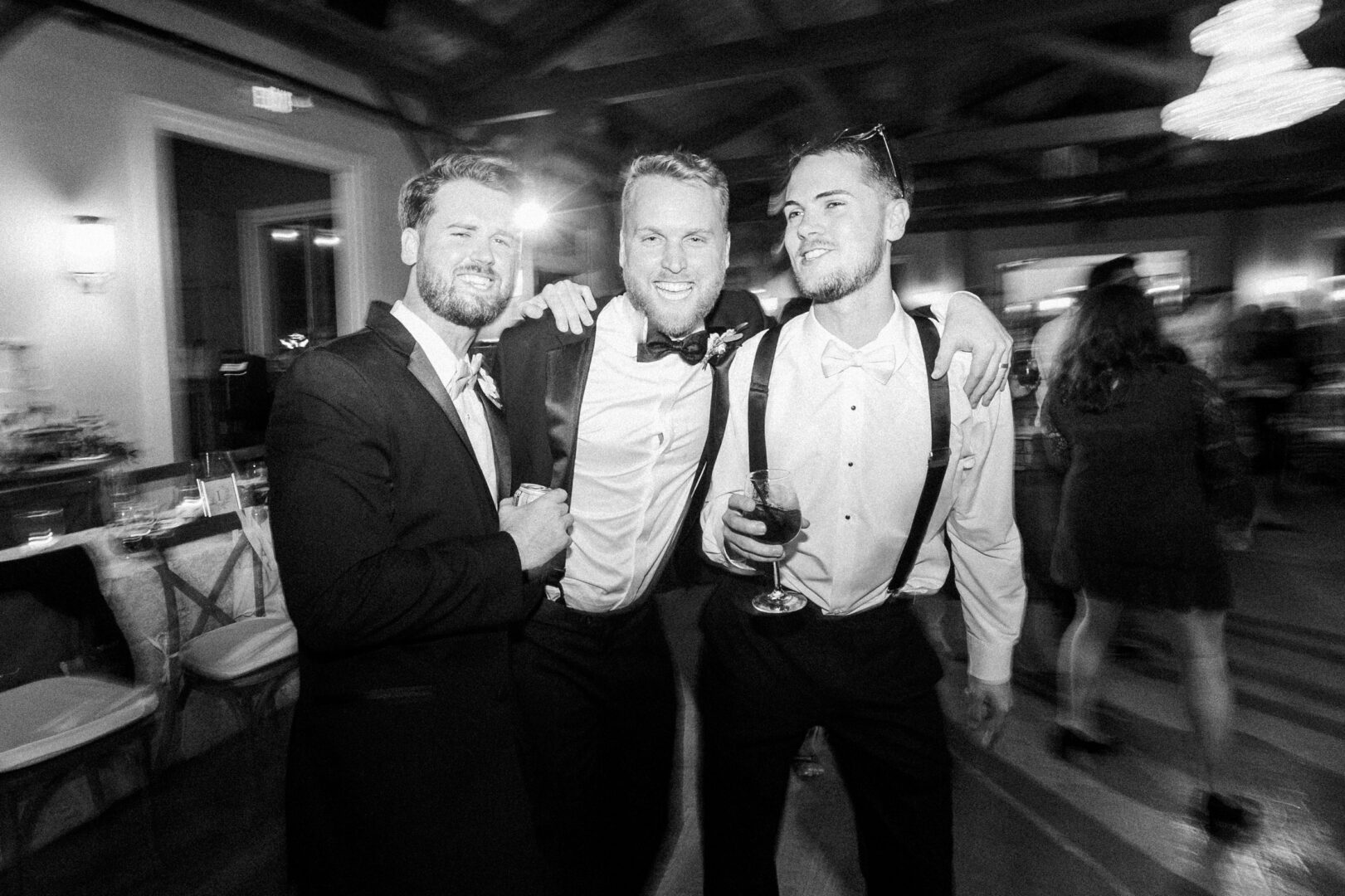 Three men in formal attire smiling and posing together at what appears to be a Willow Creek Winery wedding, with one holding a glass of wine. The background is a dimly lit room filled with guests and elegantly set tables.