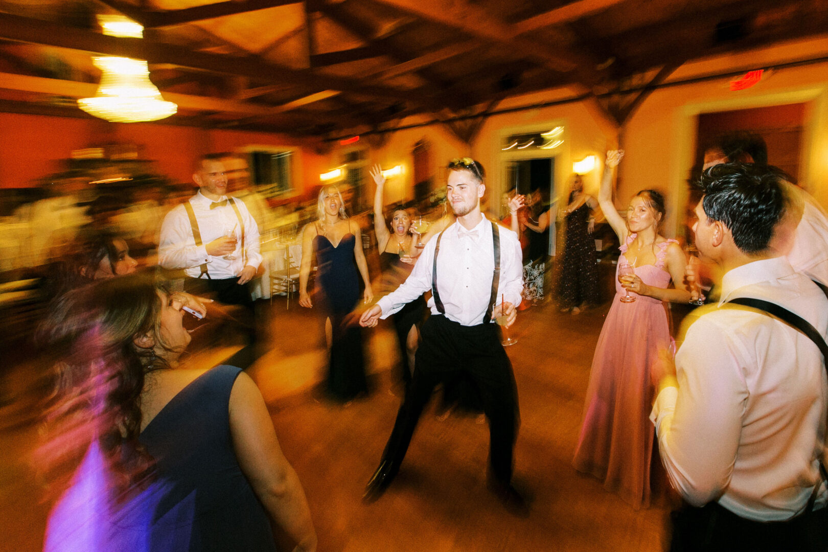 A lively group dances energetically at a Willow Creek Winery wedding, surrounded by the warm glow of wooden beams and a chandelier above.