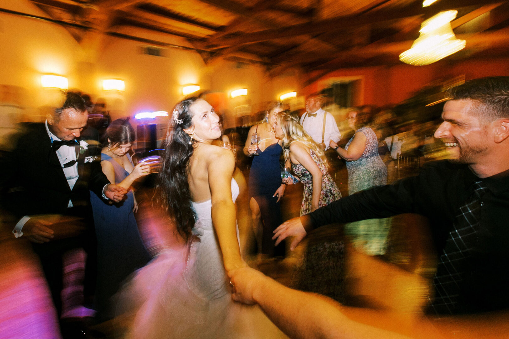 At a Willow Creek Winery wedding, people dance energetically in the warmly lit room, holding hands and smiling, with motion blur capturing their joyous movement.