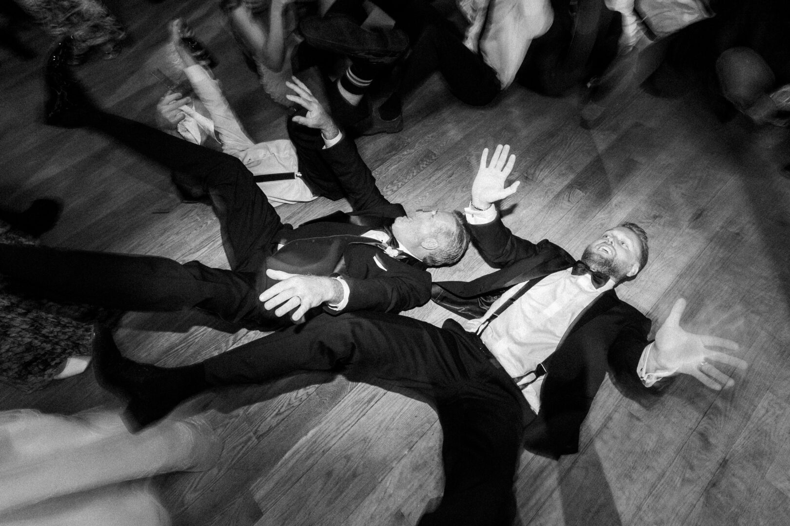 Two men in suits lie on their backs on the wooden floor at a Willow Creek Winery wedding, surrounded by others. Their arms and legs are raised, and they appear to be laughing.