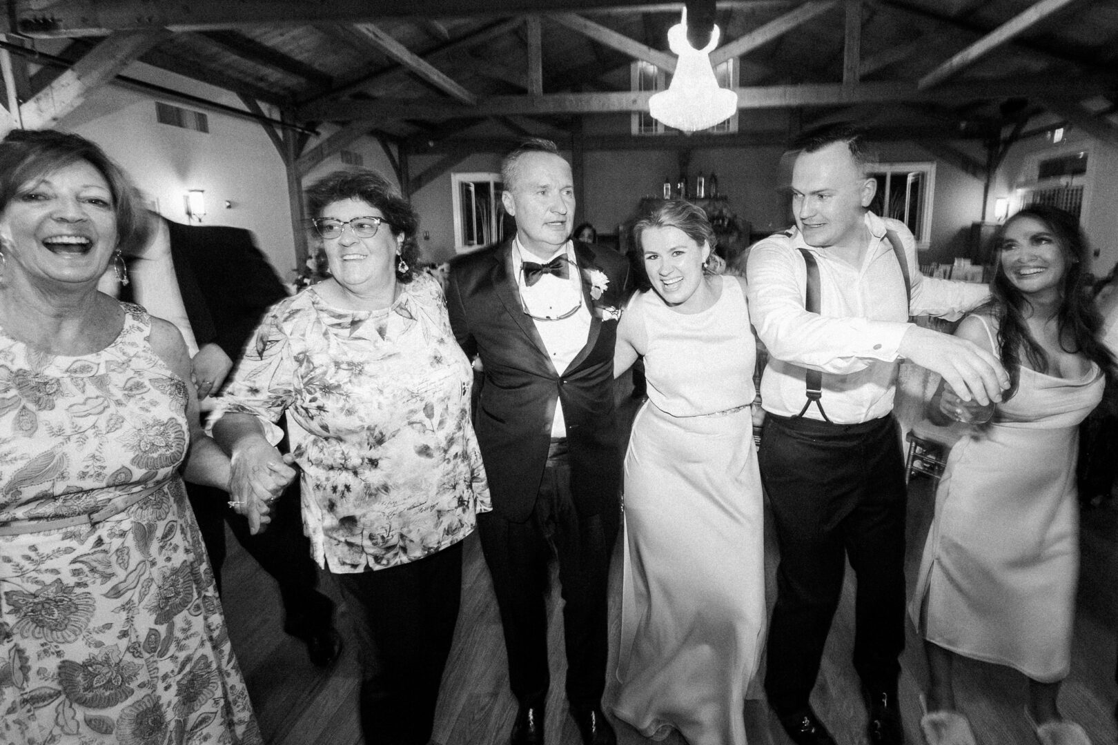 A group of people, including two men in formal attire and women in dresses, stands closely together, smiling and linking arms in the dimly lit, rustic interior setting of Willow Creek Winery.