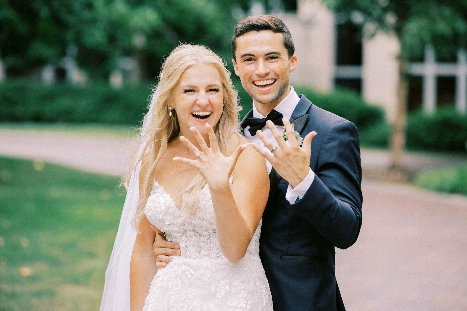 The bride and groom, glowing with happiness, proudly display their rings amidst the enchanting backdrop of a tree-lined path at their Normandy Farm wedding.