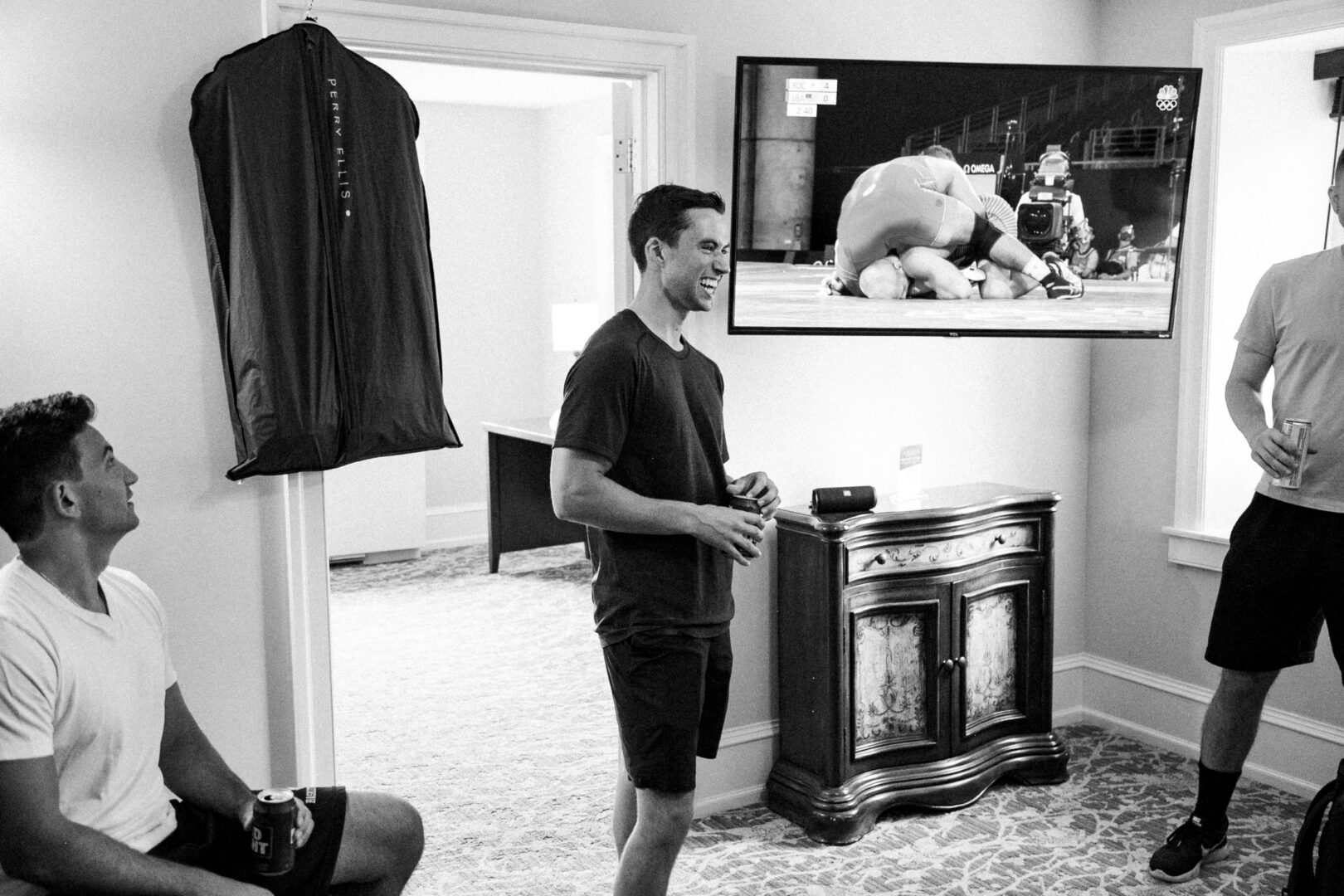 Three men in a room watch a wrestling match on TV. One man stands holding a drink, while the others sit and stand, drinks in hand. A suit bag hangs on the wall, hinting at the anticipation of an upcoming event, perhaps at Normandy Farm Wedding.