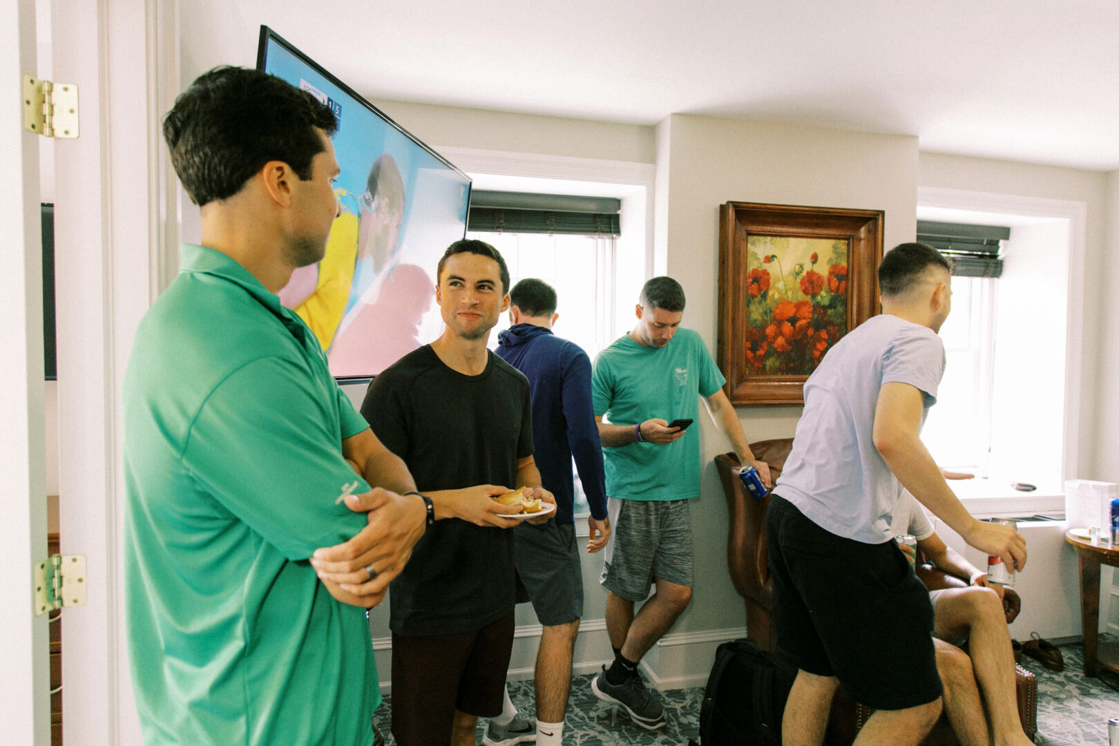 In a cozy room reminiscent of a Normandy Farm wedding, several people gather. Some are engaged in conversation while others glance at their phones. A television shares the wall with an elegant painting, completing the scene.