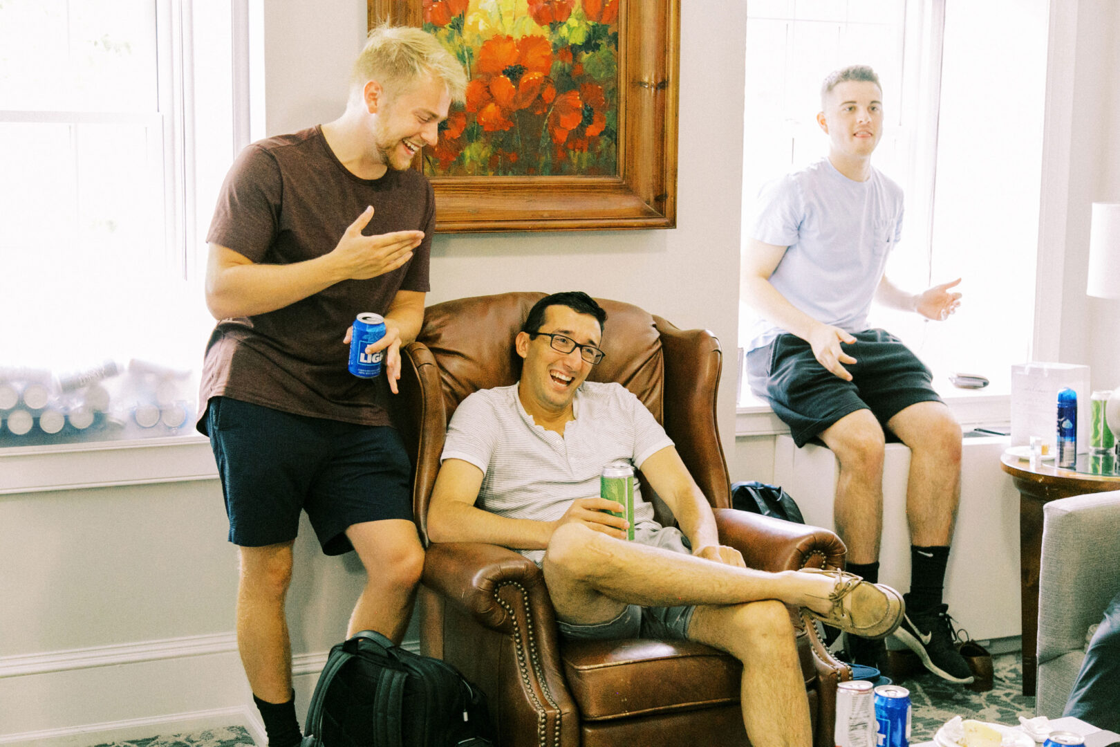 Three people relax indoors at a Normandy Farm wedding; one sits in a leather chair holding a can, while the other two stand nearby with drinks. A colorful painting hangs on the wall, adding to the celebratory atmosphere.