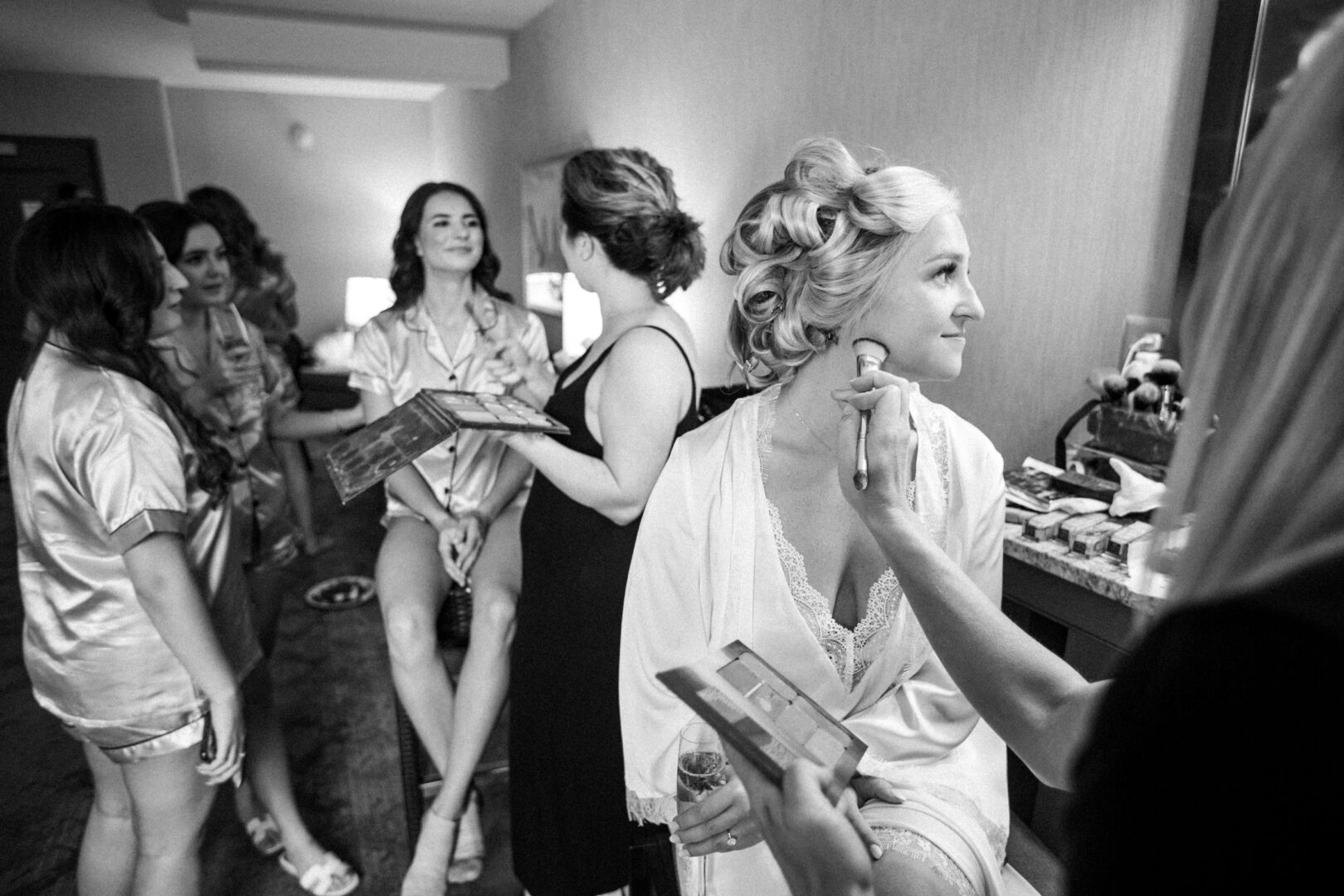 In a charming room at Normandy Farm, a group of women are getting ready; some are sitting while others elegantly apply makeup, preparing for the wedding celebration.