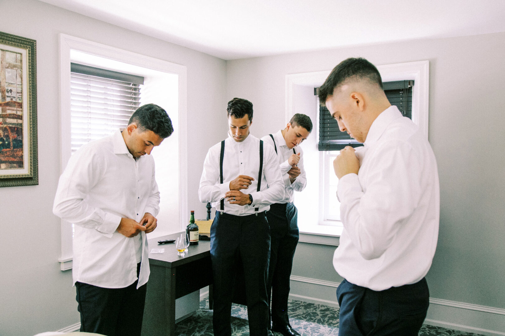 Four men in dress shirts and suspenders are adjusting their attire in a well-lit room with two windows, preparing for the Normandy Farm wedding.