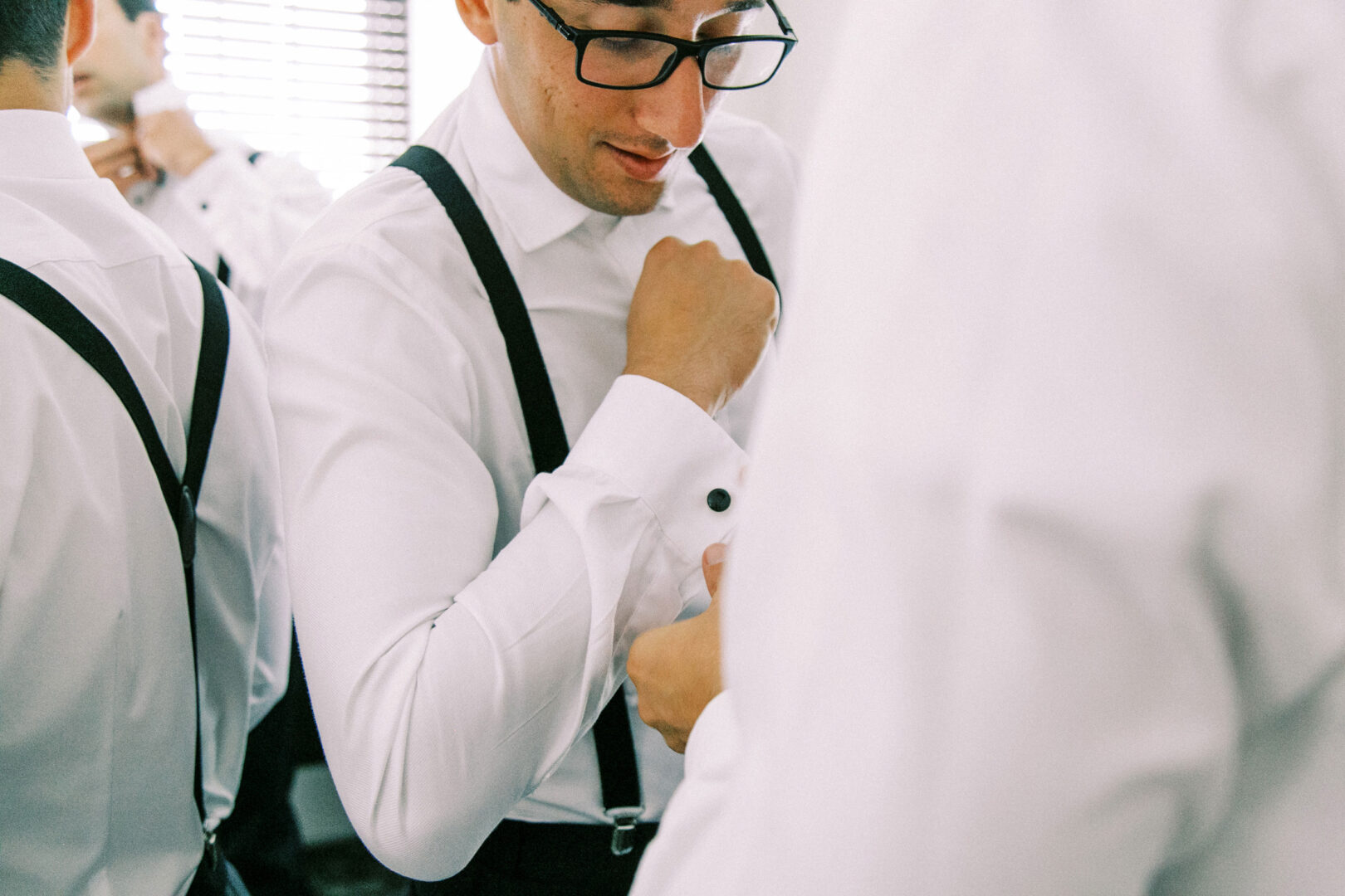 A man in glasses adjusts the cuff of his white shirt, wearing suspenders, as he stands at a Normandy Farm wedding, surrounded by others preparing in similar attire.