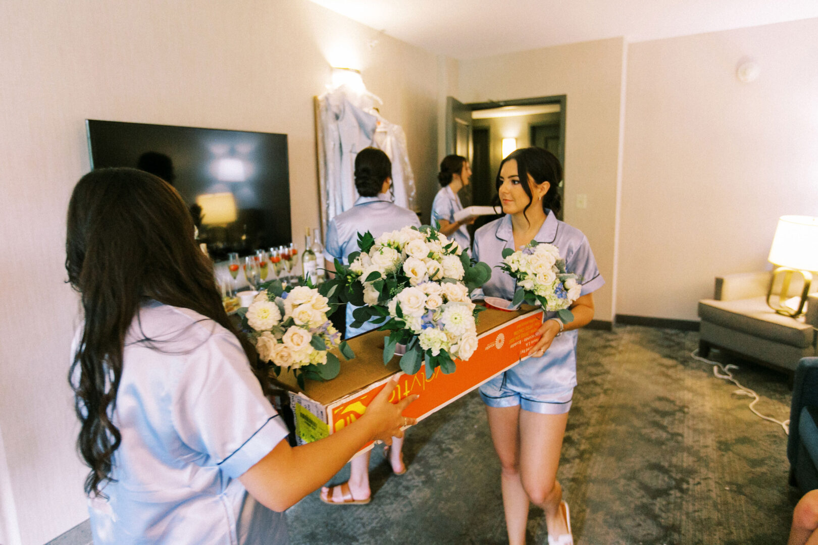 Two women in matching outfits carry a large box filled with white flowers, setting the scene for a Normandy Farm wedding in a cozy room with a TV and couch.