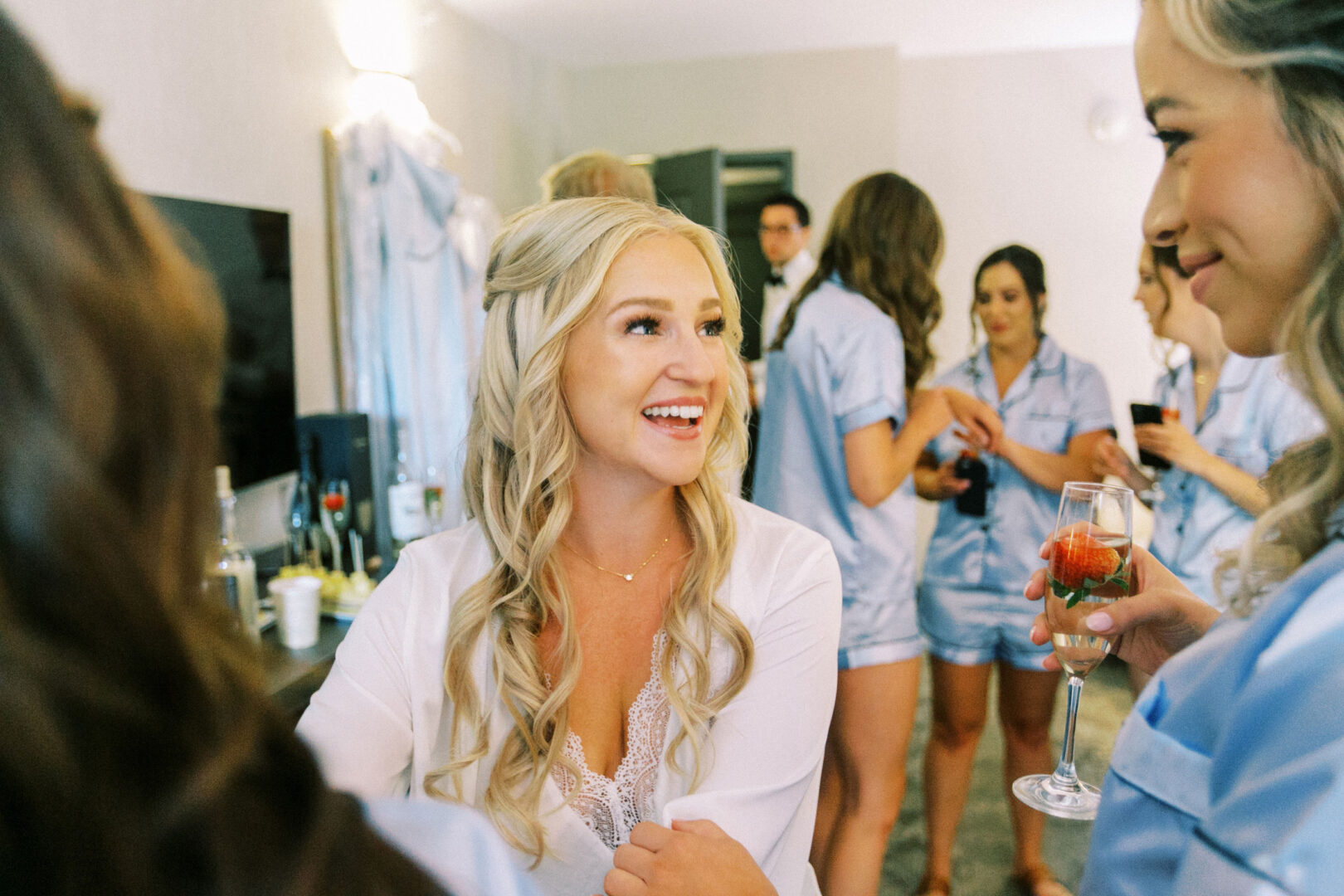 A smiling woman in a white outfit, perhaps at a Normandy Farm Wedding, is surrounded by friends in blue outfits, holding drinks, in a lively indoor setting.