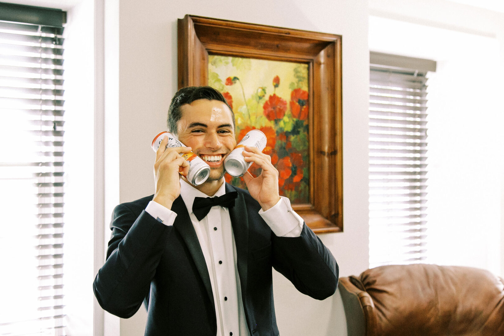 Man in a tuxedo, smiling exuberantly as he holds four cans near his face, conjures the joy of a Normandy Farm Wedding. A painting with red flowers blooms vibrantly on the wall behind him, echoing the elegance of the moment.