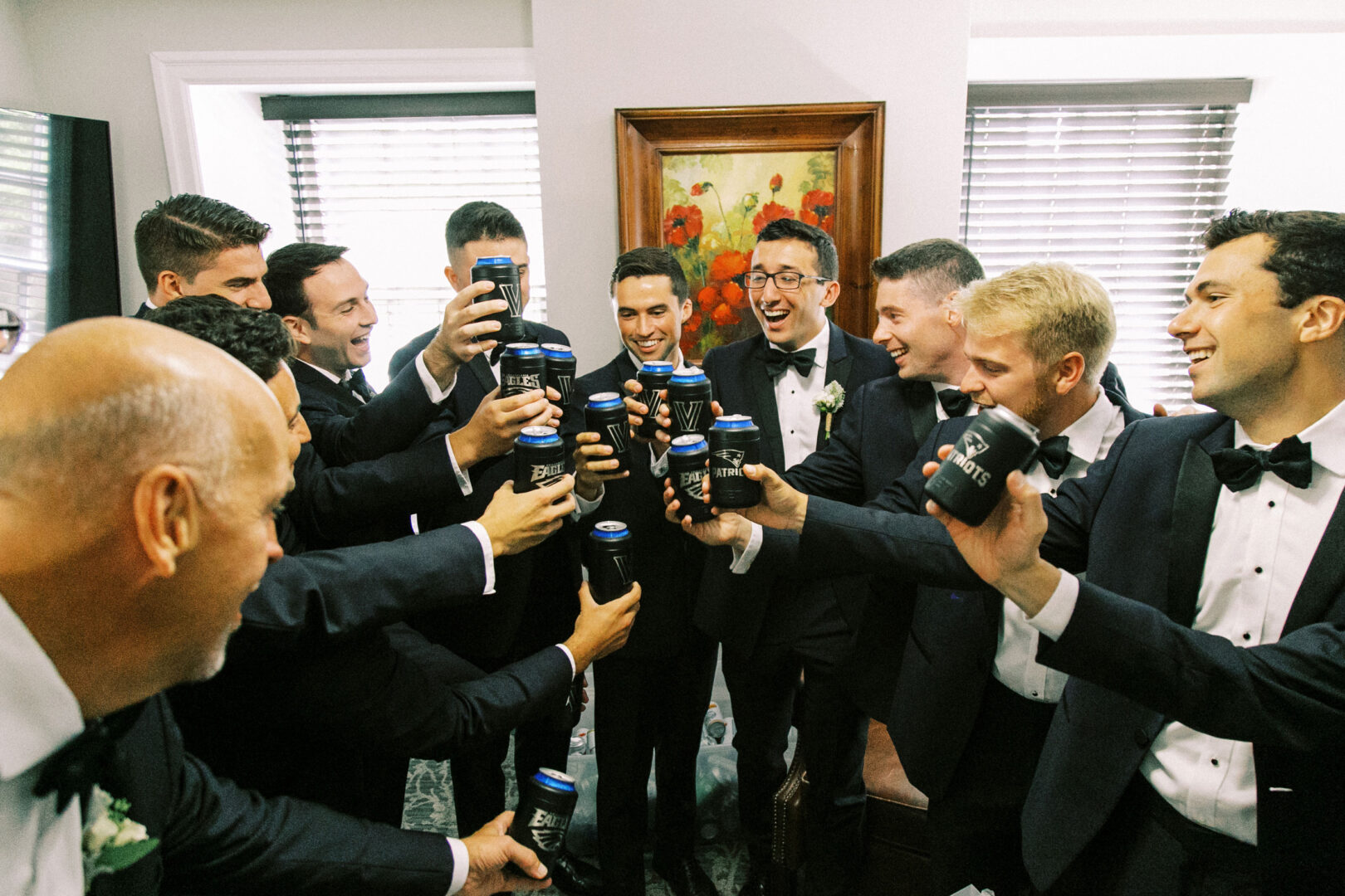 A group of men in tuxedos at the Normandy Farm Wedding toast with canned beverages indoors.