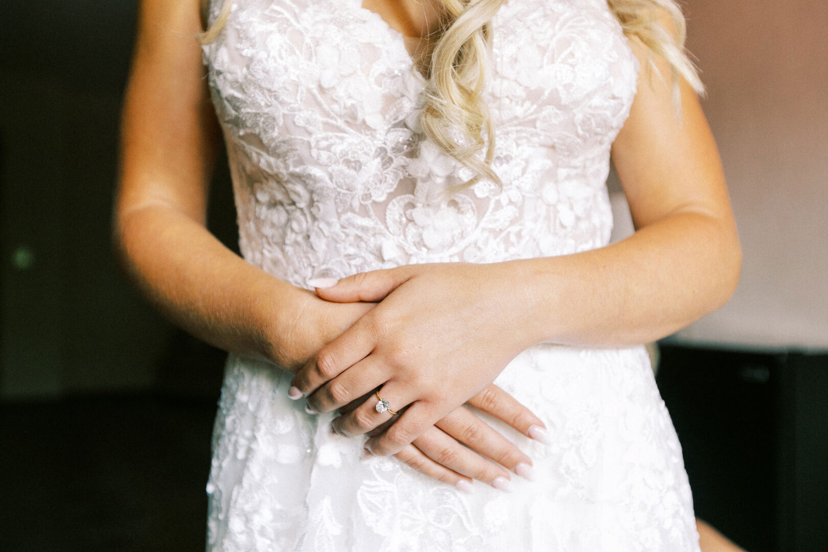 A person in a lace wedding dress stands with hands clasped, elegantly showcasing a ring, as sunlight gently filters through the rustic charm of Normandy Farm.