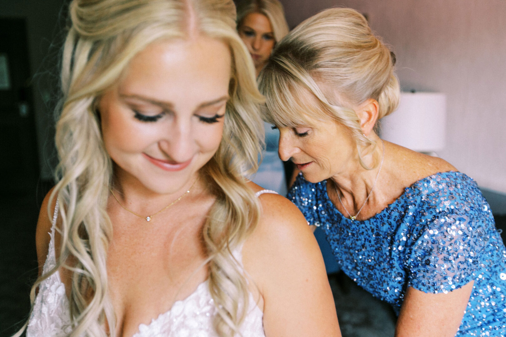 A bride smiling as a woman in a blue dress adjusts her wedding gown at the picturesque Normandy Farm Wedding venue. Another woman lingers in the background, adding to the serene atmosphere.