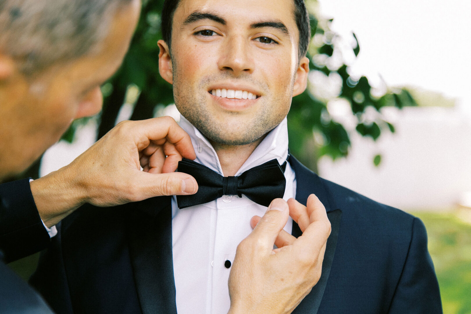 At a picturesque Normandy Farm wedding, a man in a suit smiles as another person adjusts his black bow tie, with blurred greenery providing a serene backdrop.