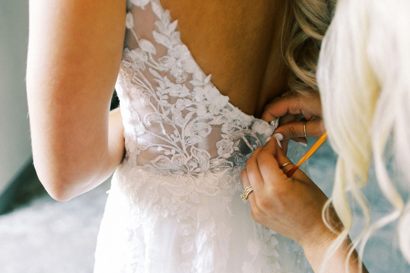 Close-up of a bride at a Normandy Farm Wedding in a lace dress being buttoned by another person, highlighting the intricate floral design on the back.