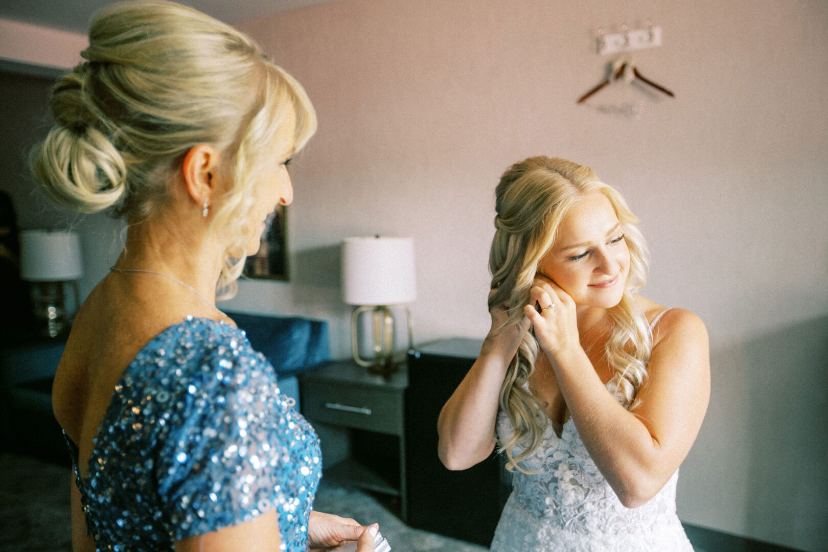 In a cozy room, two women prepare for a Normandy Farm wedding. One adjusts her earrings, while the other, clad in blue, looks on. A blue sofa and lamp provide the perfect backdrop.