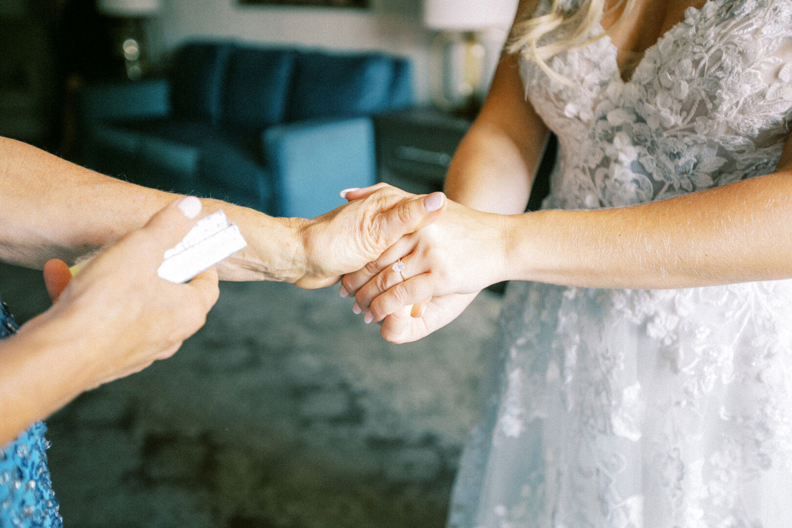 A person in a white lace dress holds hands with another in blue at a Normandy Farm Wedding. A card is visible in the hand of the person in blue, with a blue couch adding to the scene's charm.