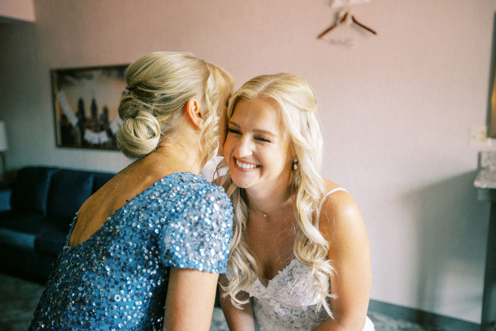 Two women smiling and hugging in a warmly lit room, one wearing a blue dress and the other in a white dress with long, wavy blonde hair, capturing the joy of their Normandy Farm wedding.