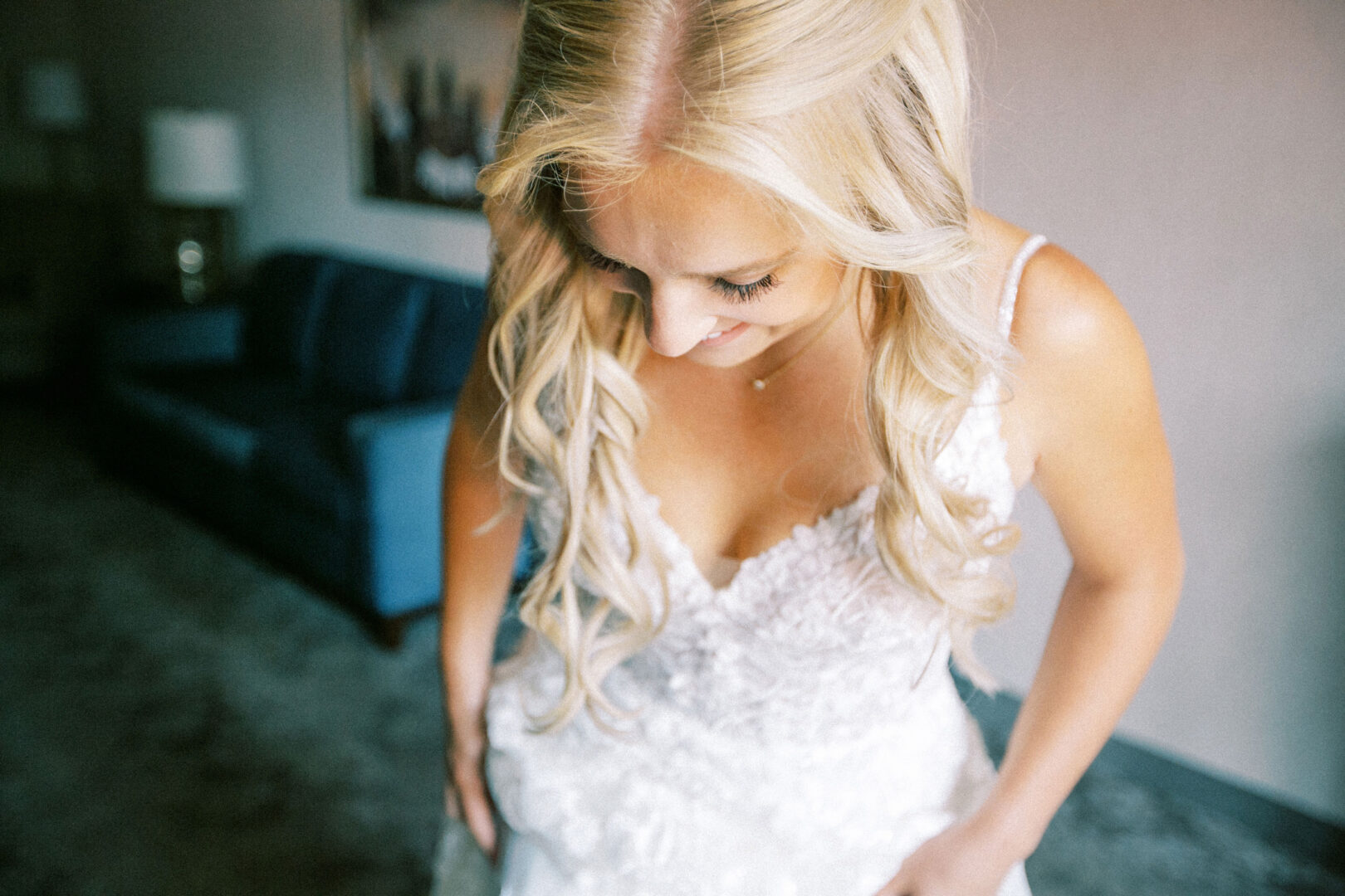 A woman in a white lace dress adjusts her outfit, preparing for her Normandy Farm wedding, with a blue sofa adding charm to the room's decor.