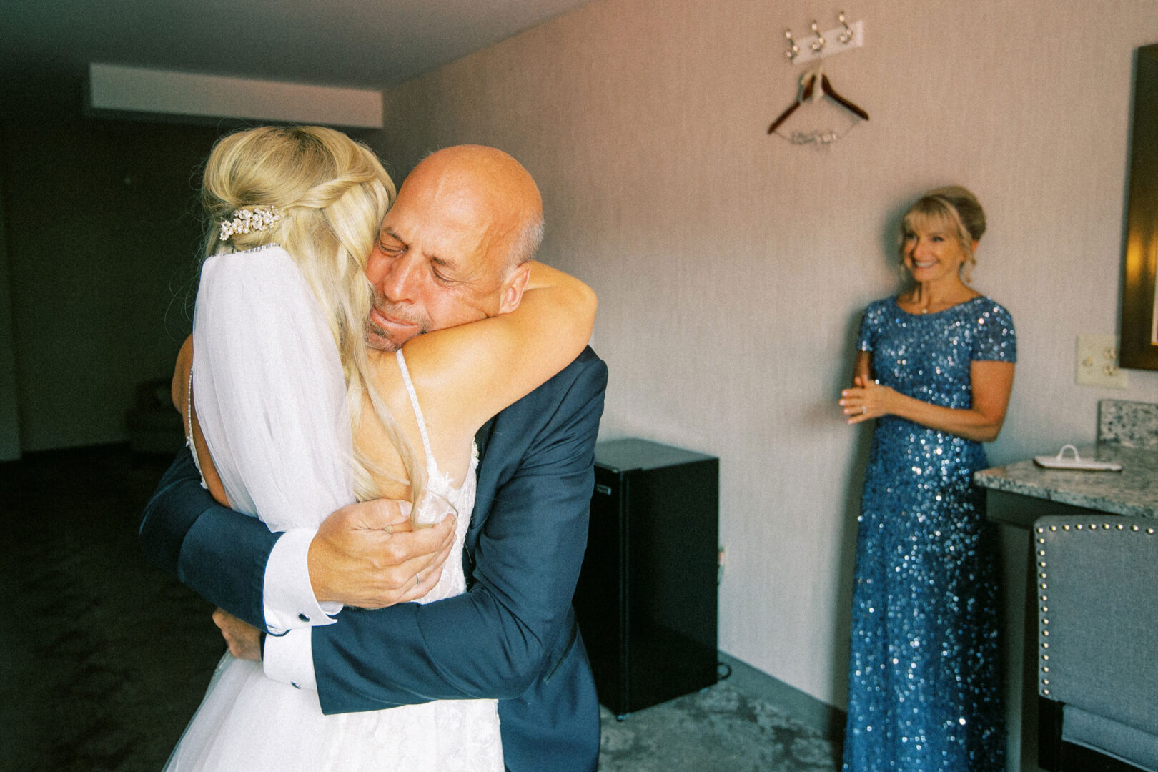 In a charming room at the Normandy Farm Wedding, a bride in a white dress embraces an older man in a suit while a woman in a blue dress stands nearby, watching them with a warm smile.