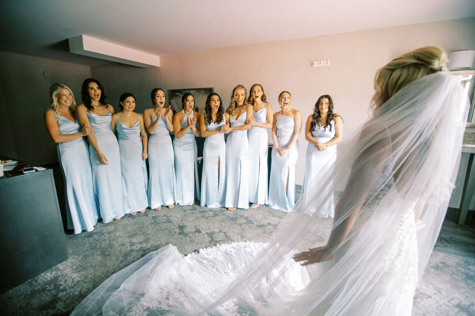 In a sunlit room at Normandy Farm Wedding, a bride in a white dress faces her bridesmaids in matching blue dresses, all smiling and bubbling with excitement.