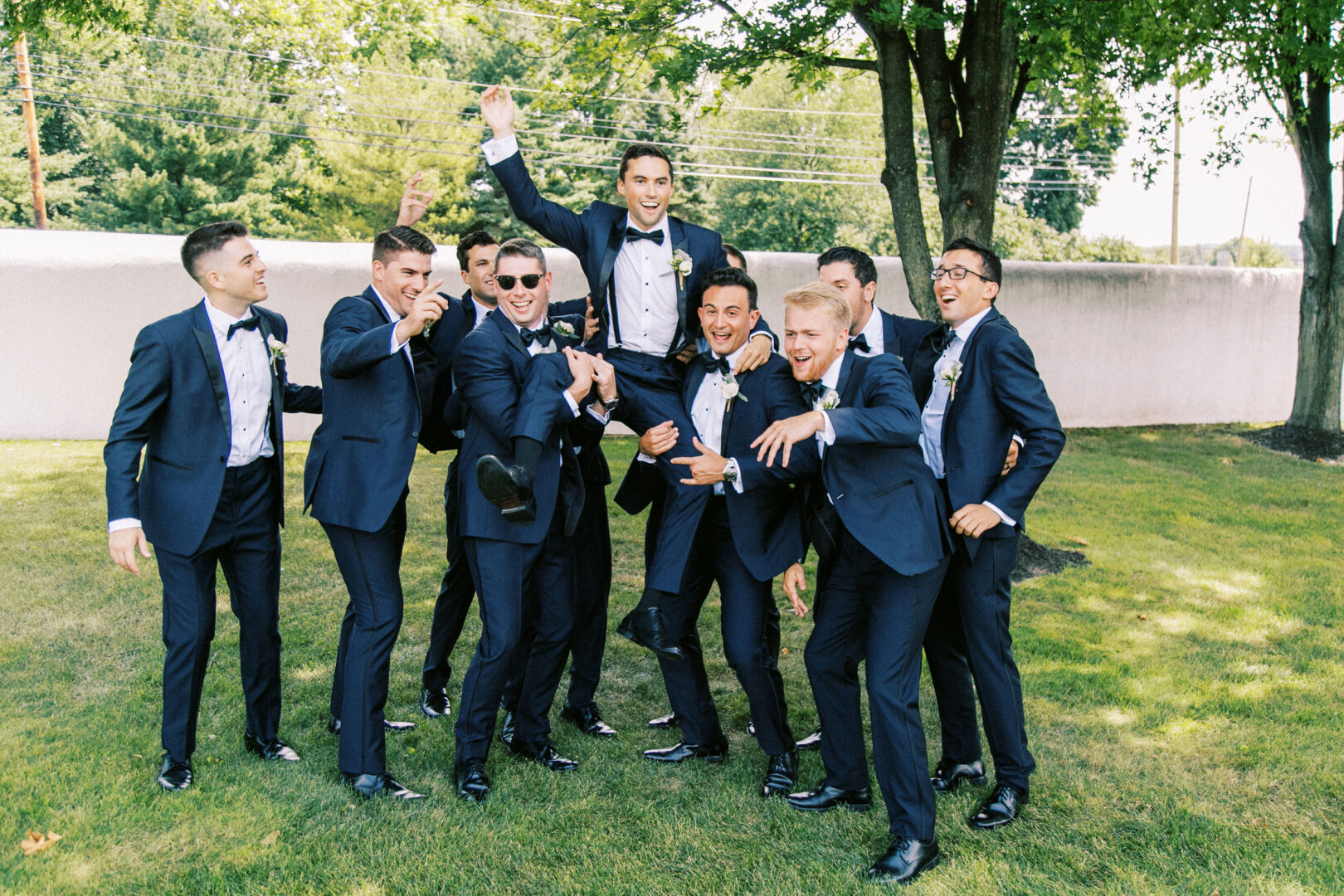 At a Normandy Farm Wedding, a group of men in suits joyfully lifts the groom, who sports sunglasses, in a grassy outdoor area.