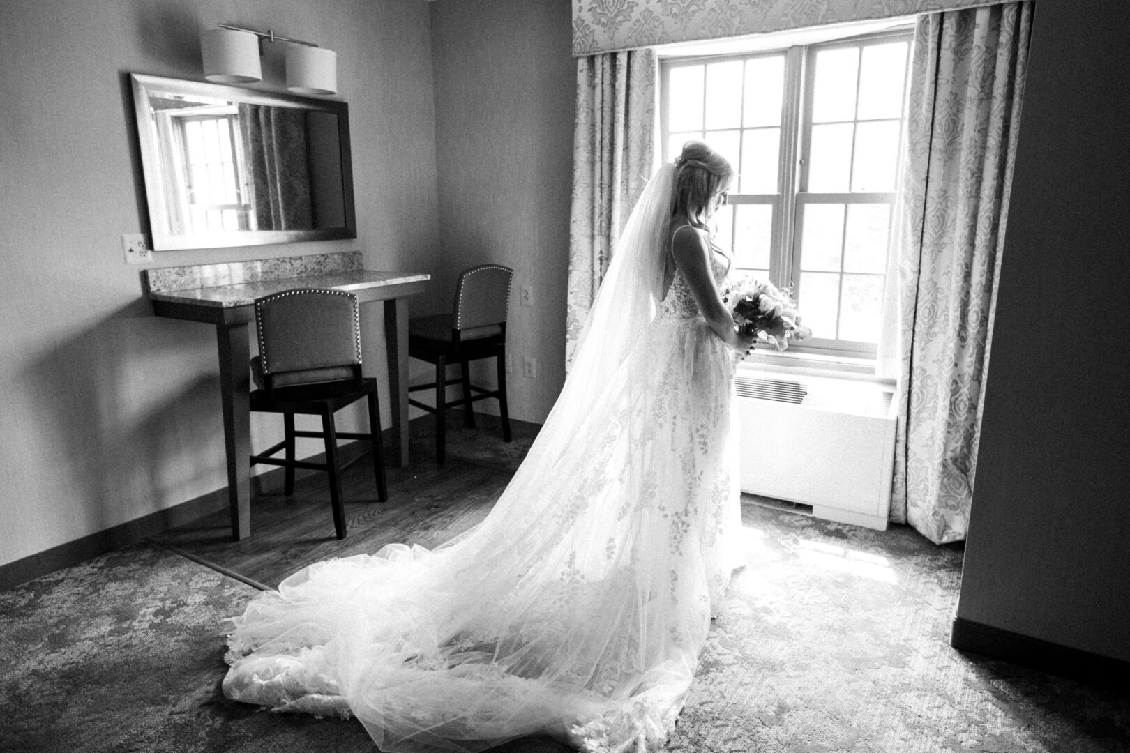 Bride in a long gown stands by a window at Normandy Farm Wedding, holding a bouquet with light streaming in. Room includes a table, chairs, and a large mirror.