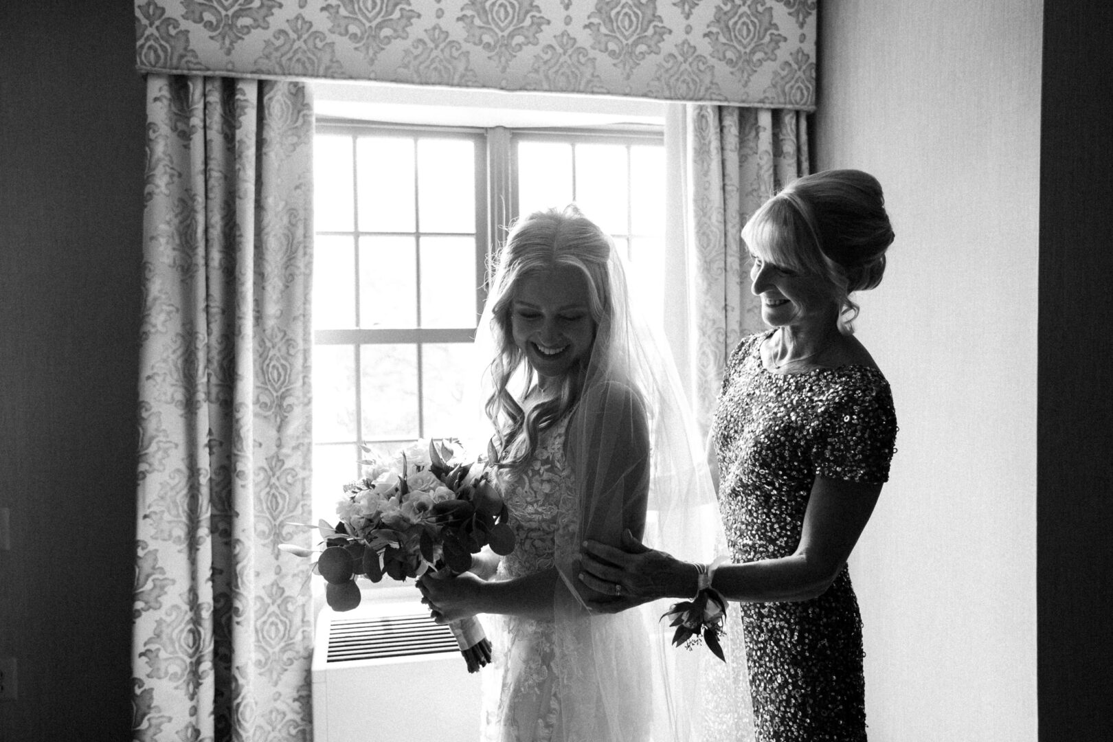 Bride holding a bouquet stands next to a woman in a sequined dress, both beaming amidst the elegant charm of a Normandy Farm Wedding, near a window with patterned curtains.