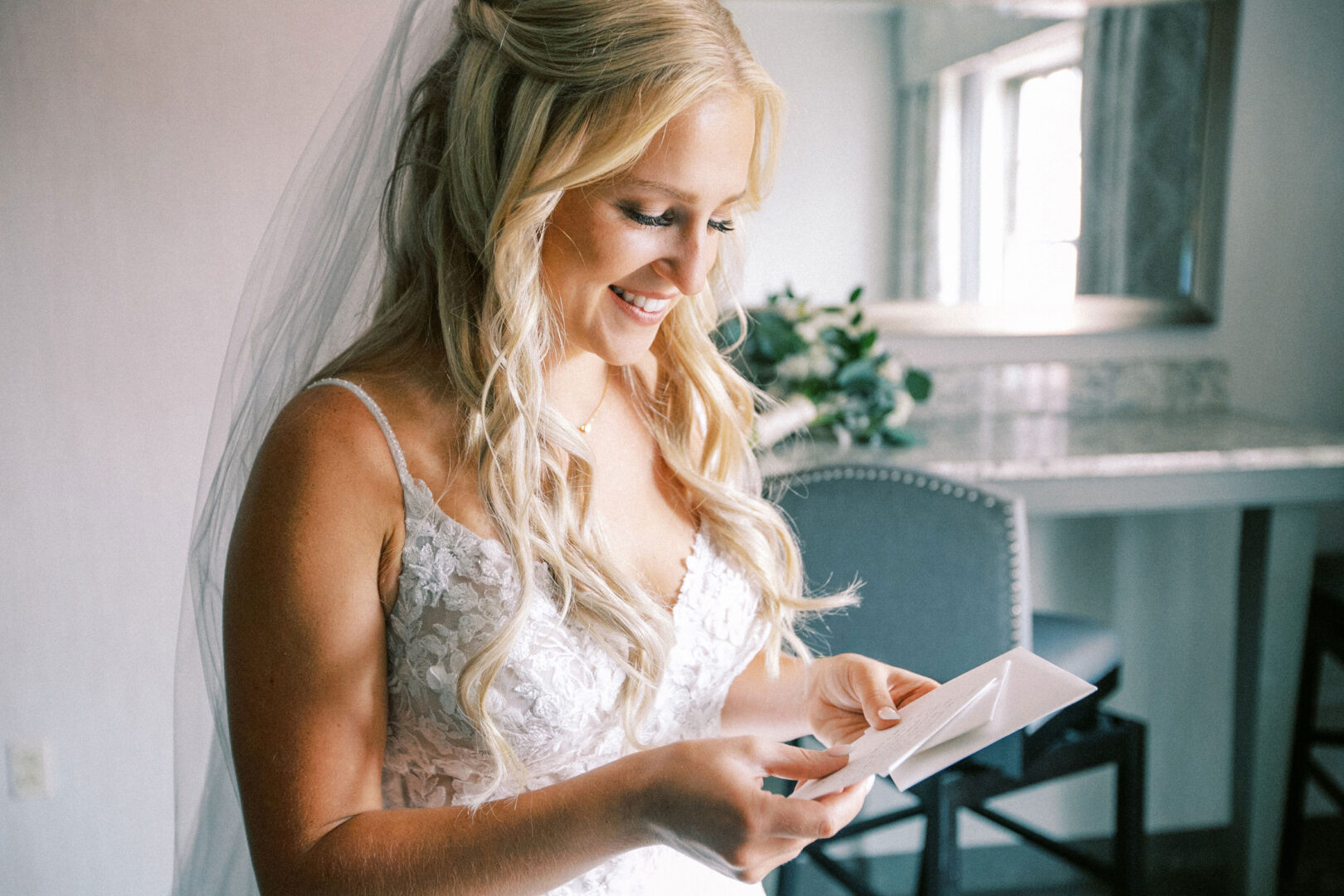 At her Normandy Farm wedding, a bride with long blonde hair in a white lace dress smiles while reading a card, surrounded by greenery and the reflection in the room's mirror.