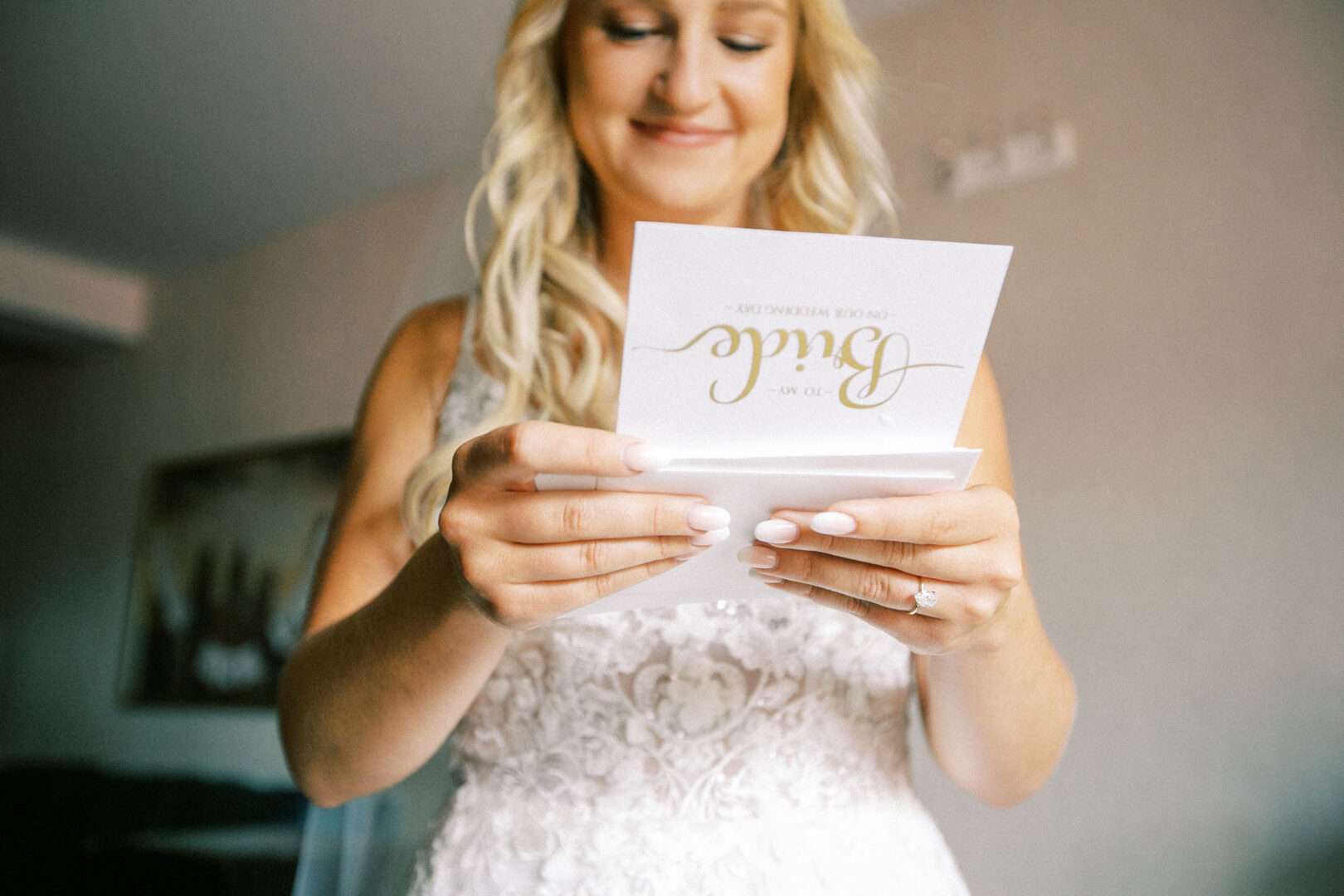 At her Normandy Farm wedding, the bride in a white lace dress smiles warmly as she reads a card inscribed with "Bride.