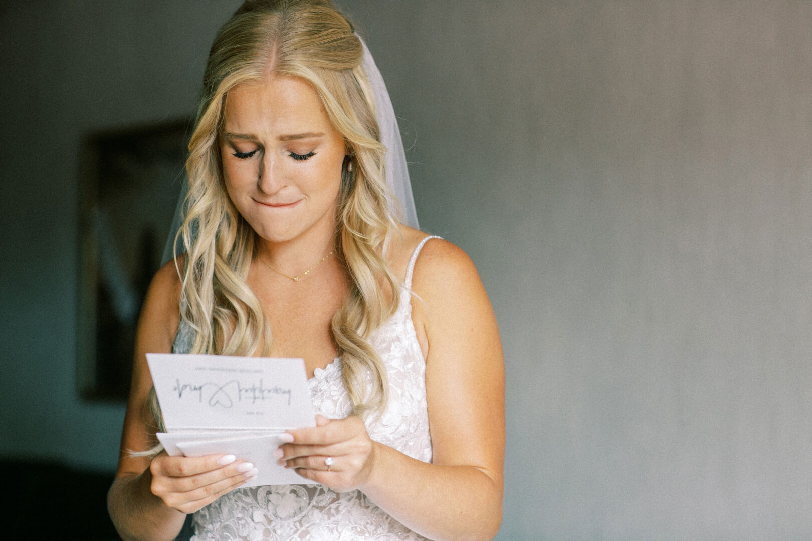 At a picturesque Normandy Farm wedding, a bride with long blonde hair and a veil stands indoors in her white dress, reading an emotional letter.