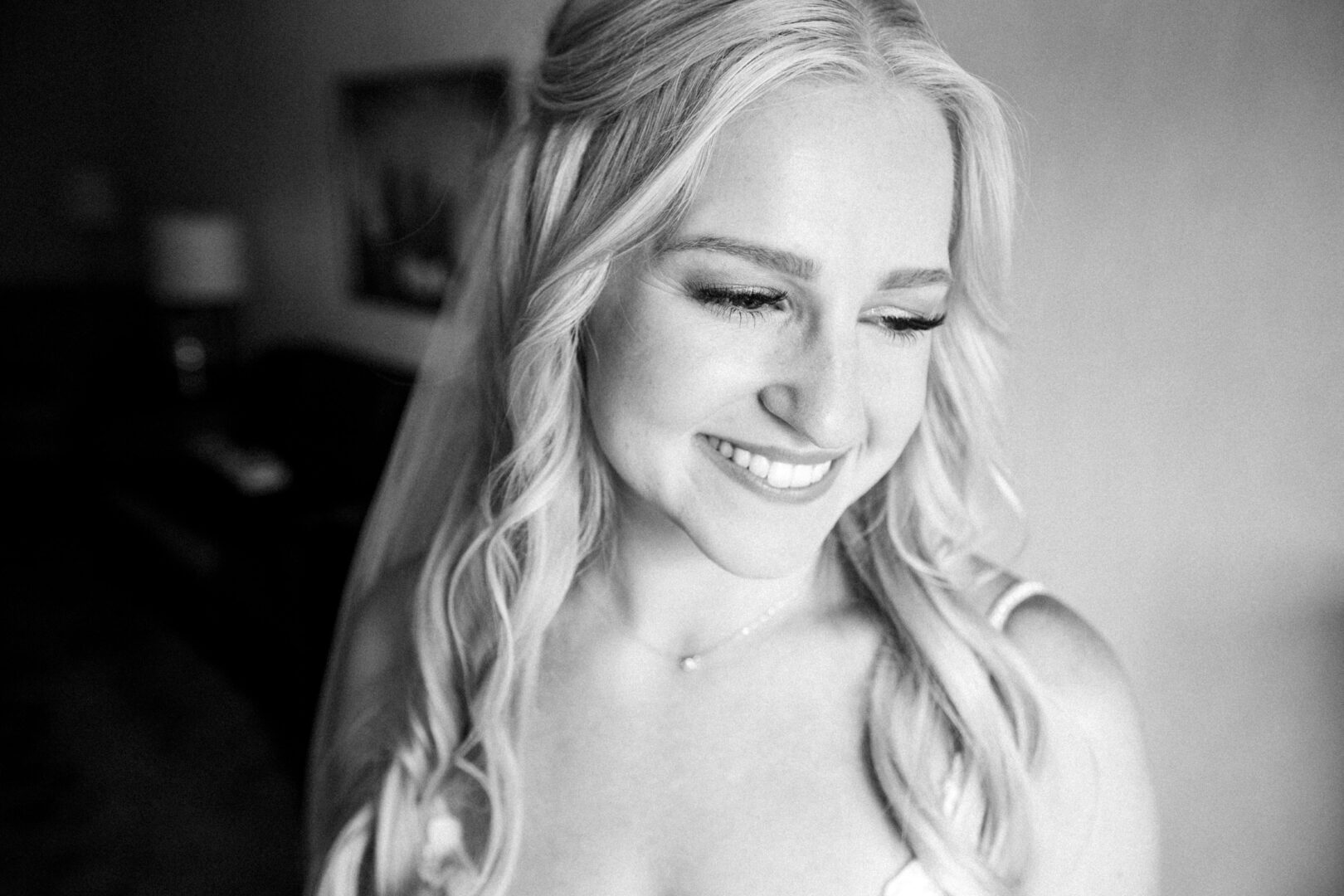 Black and white photo of a smiling person with long, wavy hair, wearing a white top. The softly blurred indoor setting evokes the warmth and elegance of a Normandy Farm wedding.