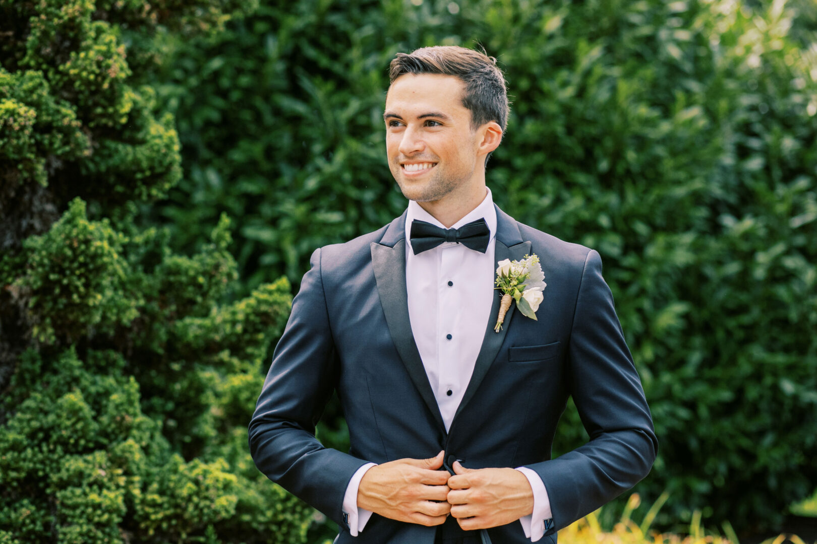 A man in a tuxedo with a bow tie smiles while adjusting his jacket amid the lush greenery of Normandy Farm. A boutonniere adorns his left lapel, adding a charming touch to this elegant wedding scene.