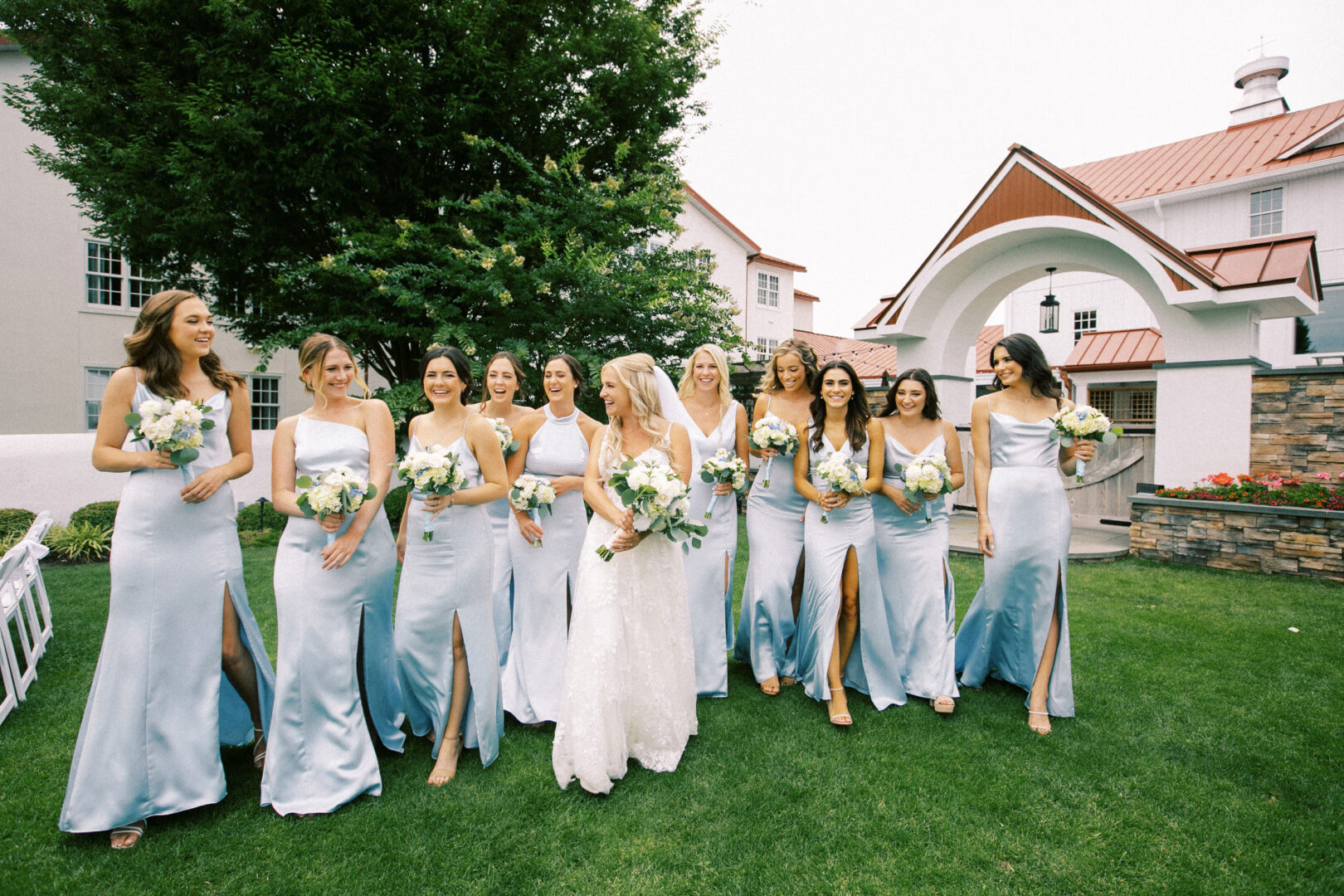 At a picturesque Normandy Farm wedding, a bride with ten bridesmaids in light blue dresses stands gracefully on the lawn. They clutch elegant bouquets of white flowers as they pose near a charming building with a red roof, capturing the essence of their special day.