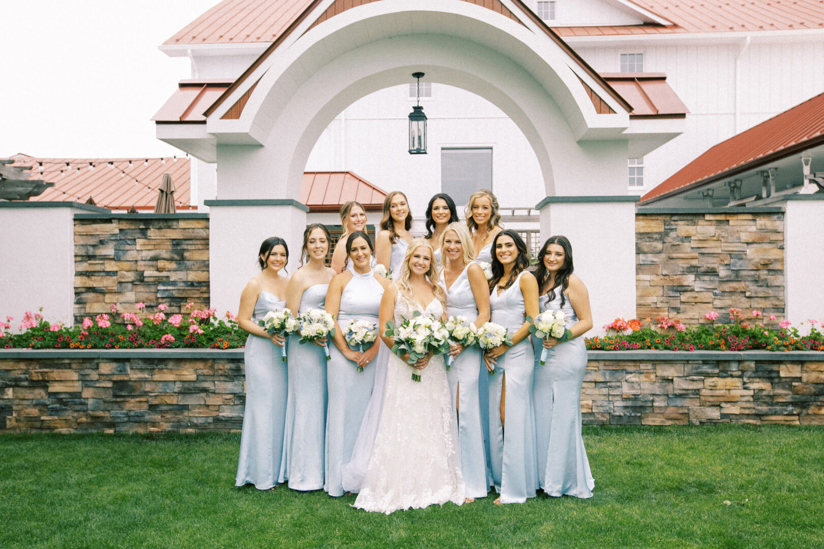 A bride in a white gown stands with her ten bridesmaids in light blue dresses, holding bouquets, in front of the charming backdrop of Normandy Farm's landscaped wall and elegant white building.