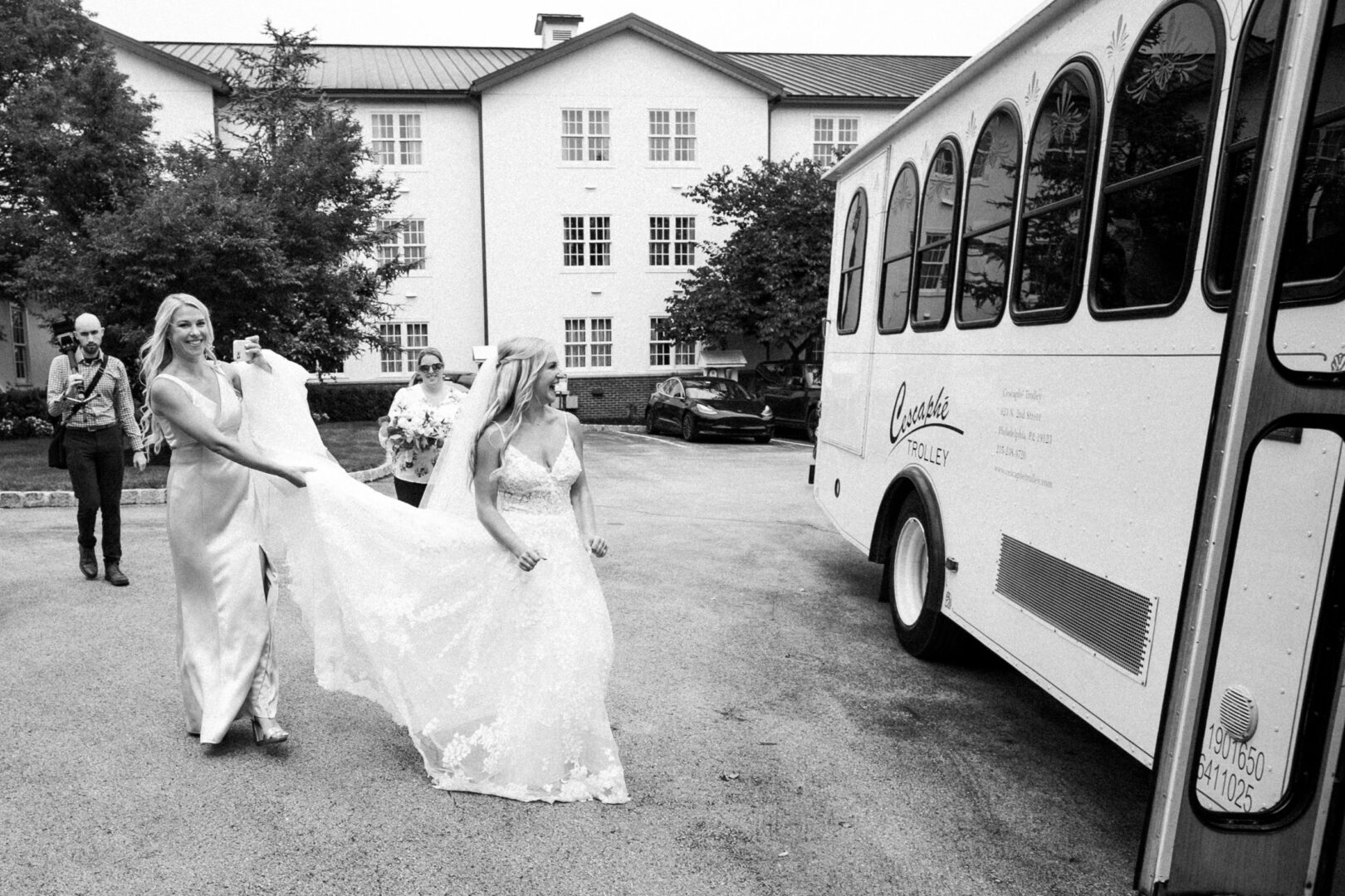 The bride gracefully walks towards the bus, her dress elegantly held by a bridesmaid, creating a picture-perfect scene in front of the charming white building, evoking the timeless beauty of a Normandy Farm Wedding.