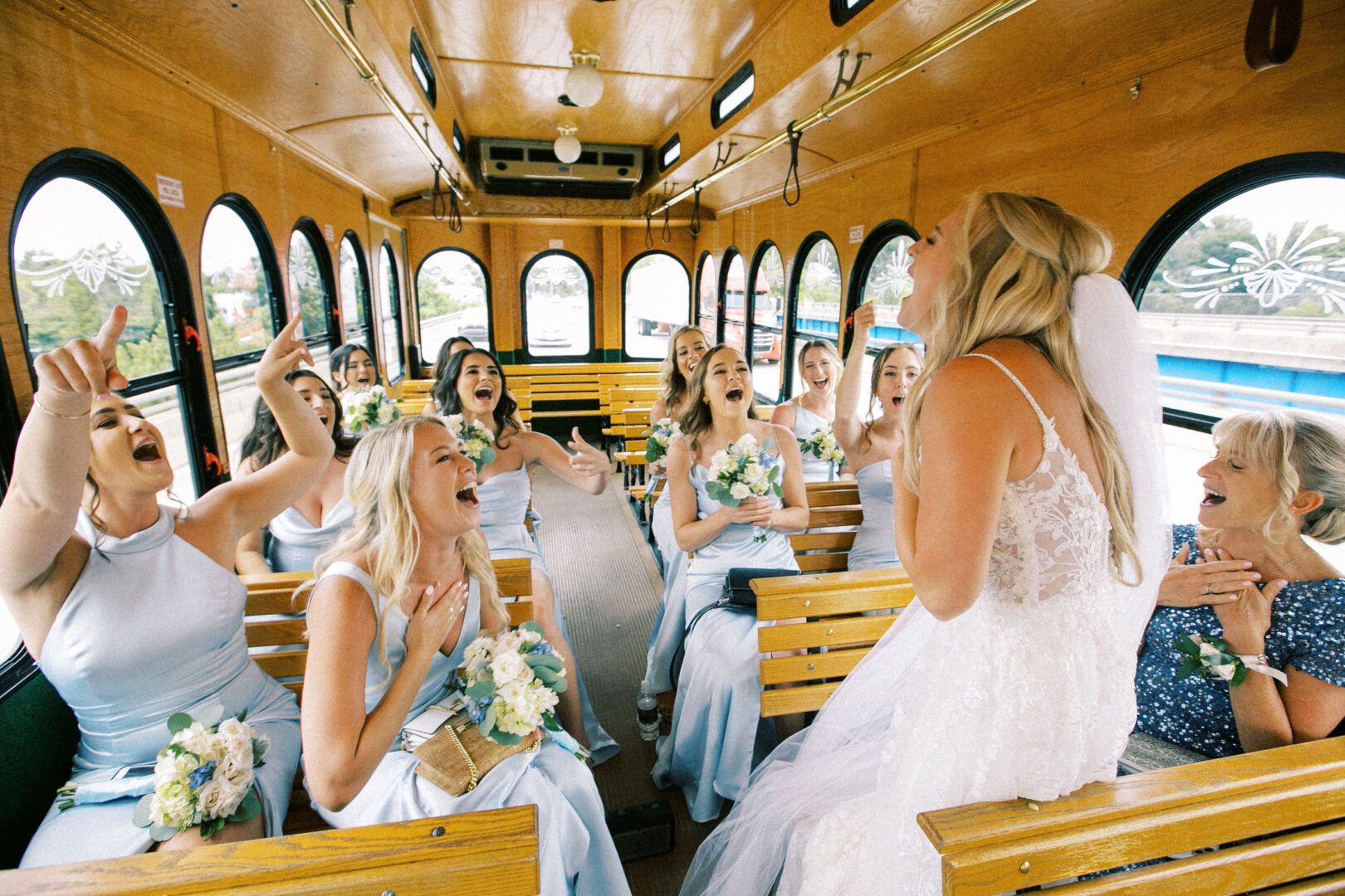 A bride in a white dress and veil celebrates her Normandy Farm wedding with bridesmaids in light blue dresses inside a wooden trolley.