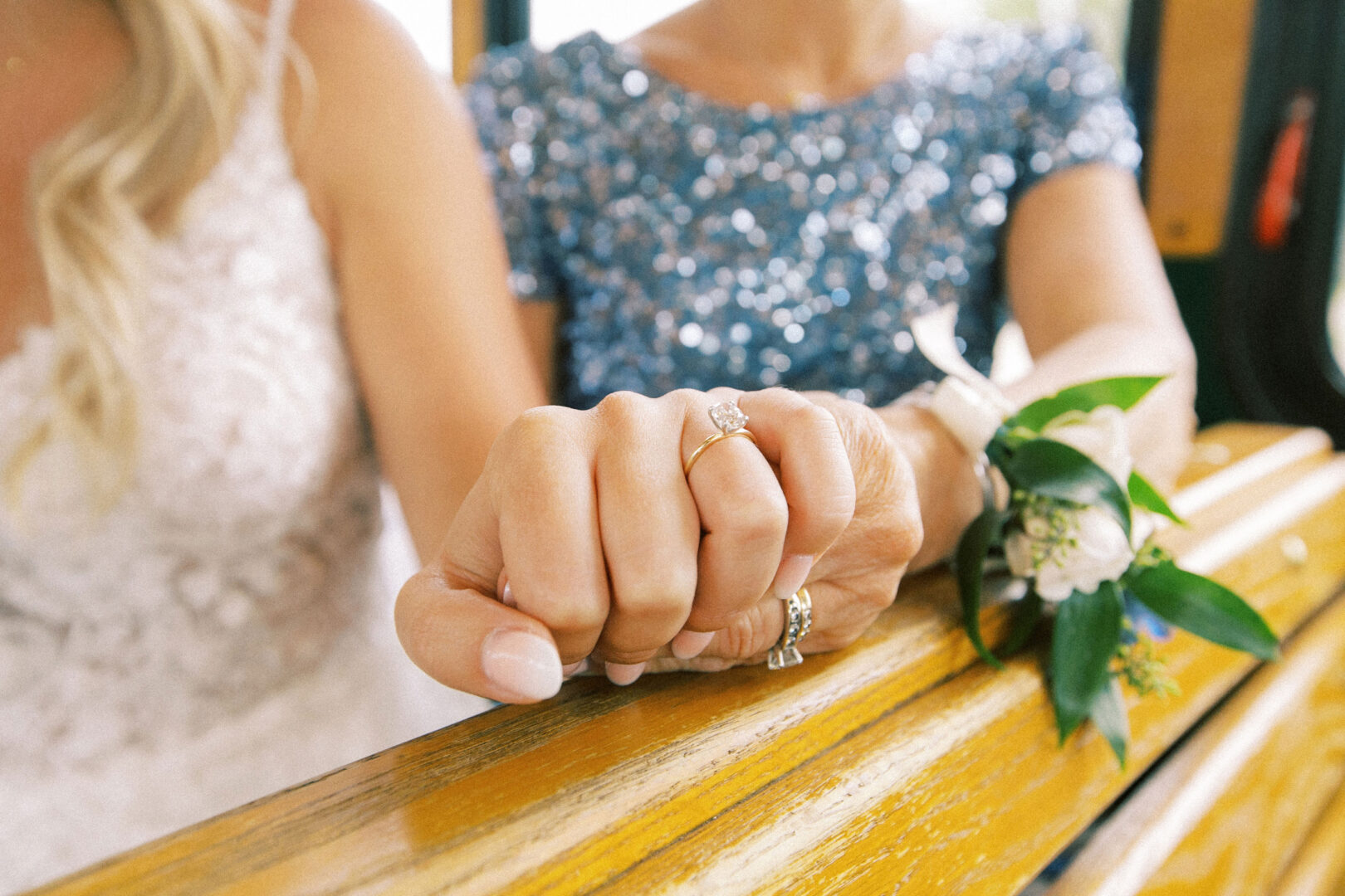 Two women display their hands, beautifully adorned with rings. One wears a white dress, the other a blue sequined one. They hold hands resting on a wooden surface, capturing the charm of a Normandy Farm Wedding.