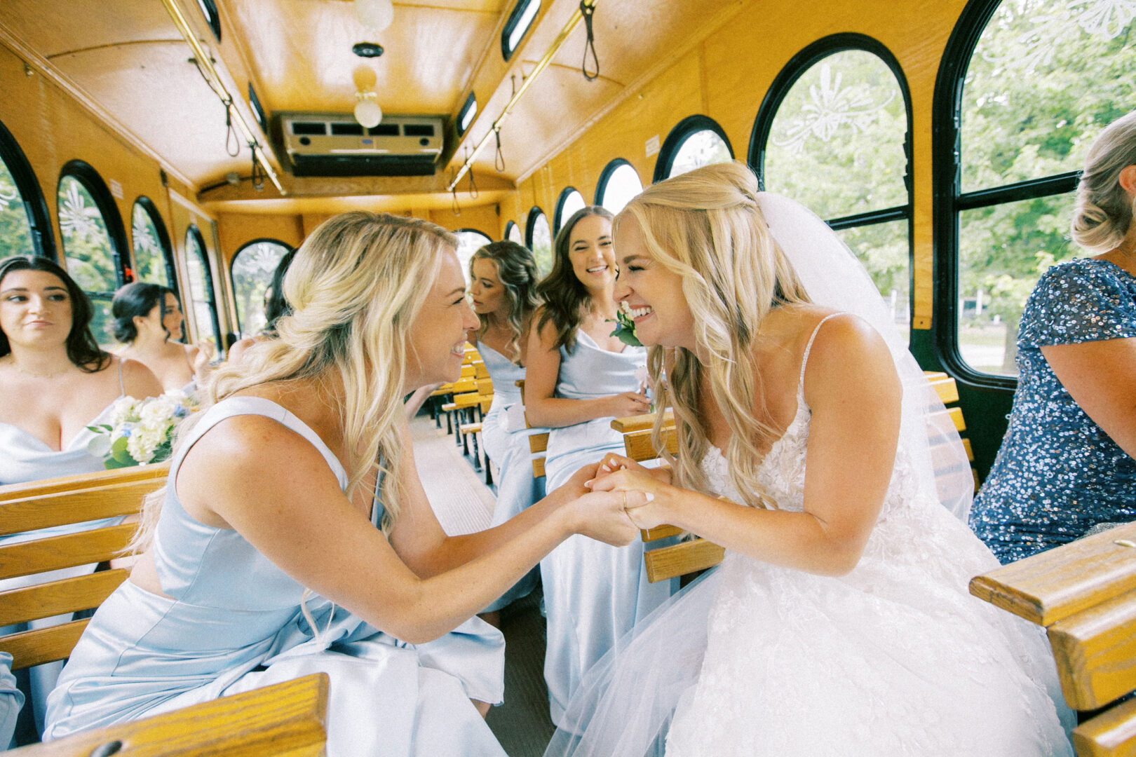 At Normandy Farm, a bride in her elegant wedding dress and a bridesmaid in blue share a joyful moment, smiling and holding hands inside a charming yellow trolley, surrounded by their fellow bridesmaids.