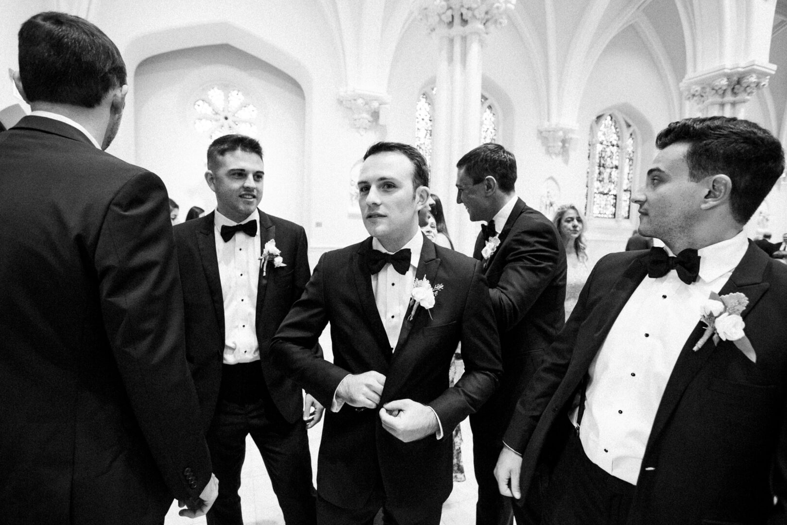 A group of five men in formal black tuxedos, adorned with boutonnières, stand indoors at a Normandy Farm wedding. The setting is an ornate room with arched windows and elegant columns.