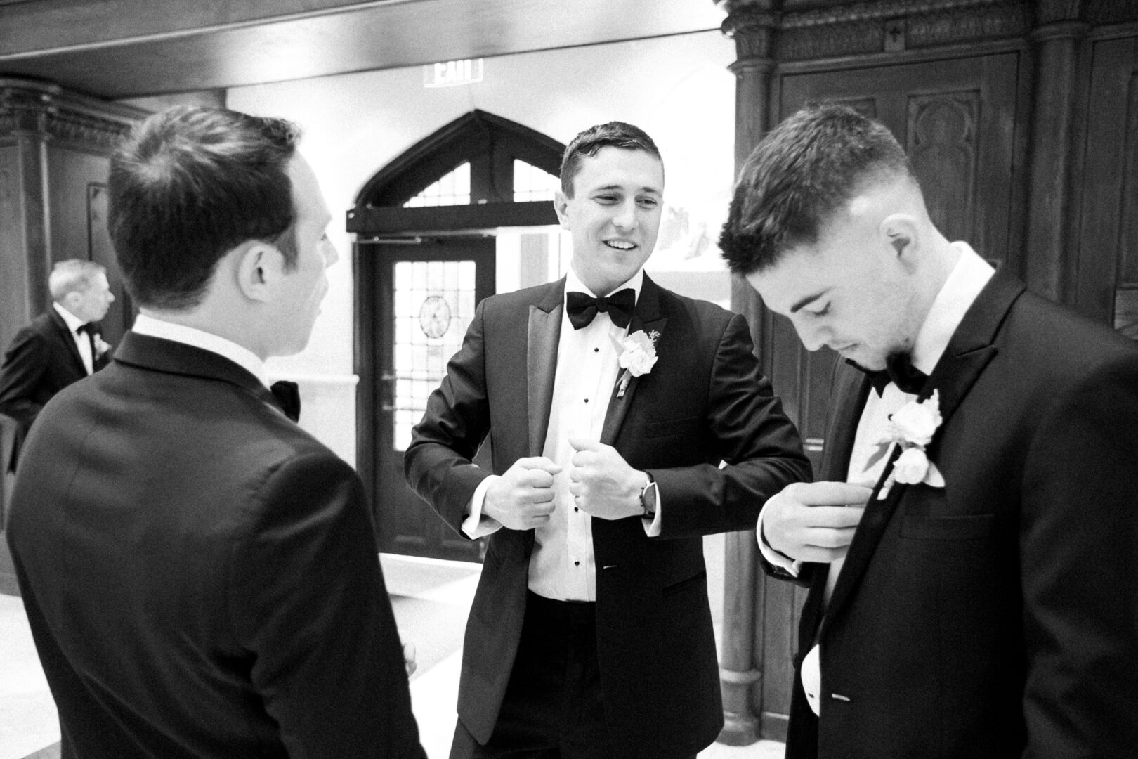 Three men in tuxedos stand in conversation indoors at the elegant Normandy Farm Wedding, adjusting their suits and boutonnieres.