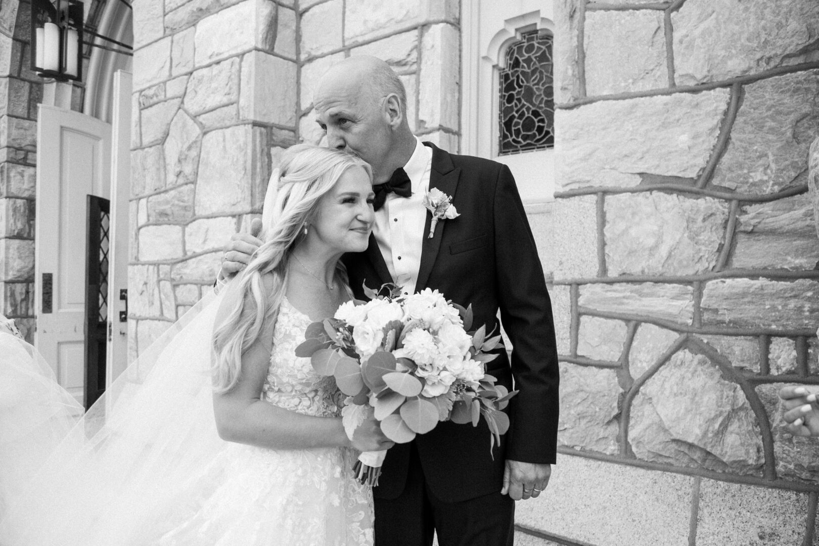 In the picturesque setting of a Normandy Farm wedding, a bride and an older man in formal attire stand outside a charming stone building. The man tenderly kisses the bride's forehead as she holds her bouquet, capturing a moment of timeless elegance and warmth.